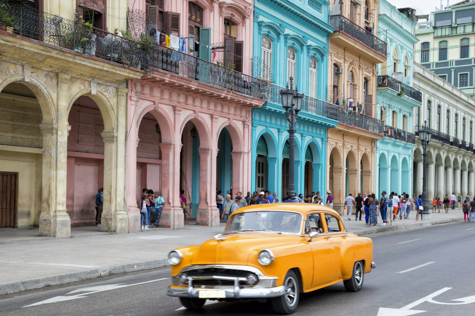 Vintage bright-yellow American car driving along brightly painted facades in Havana Vieja, Cuba