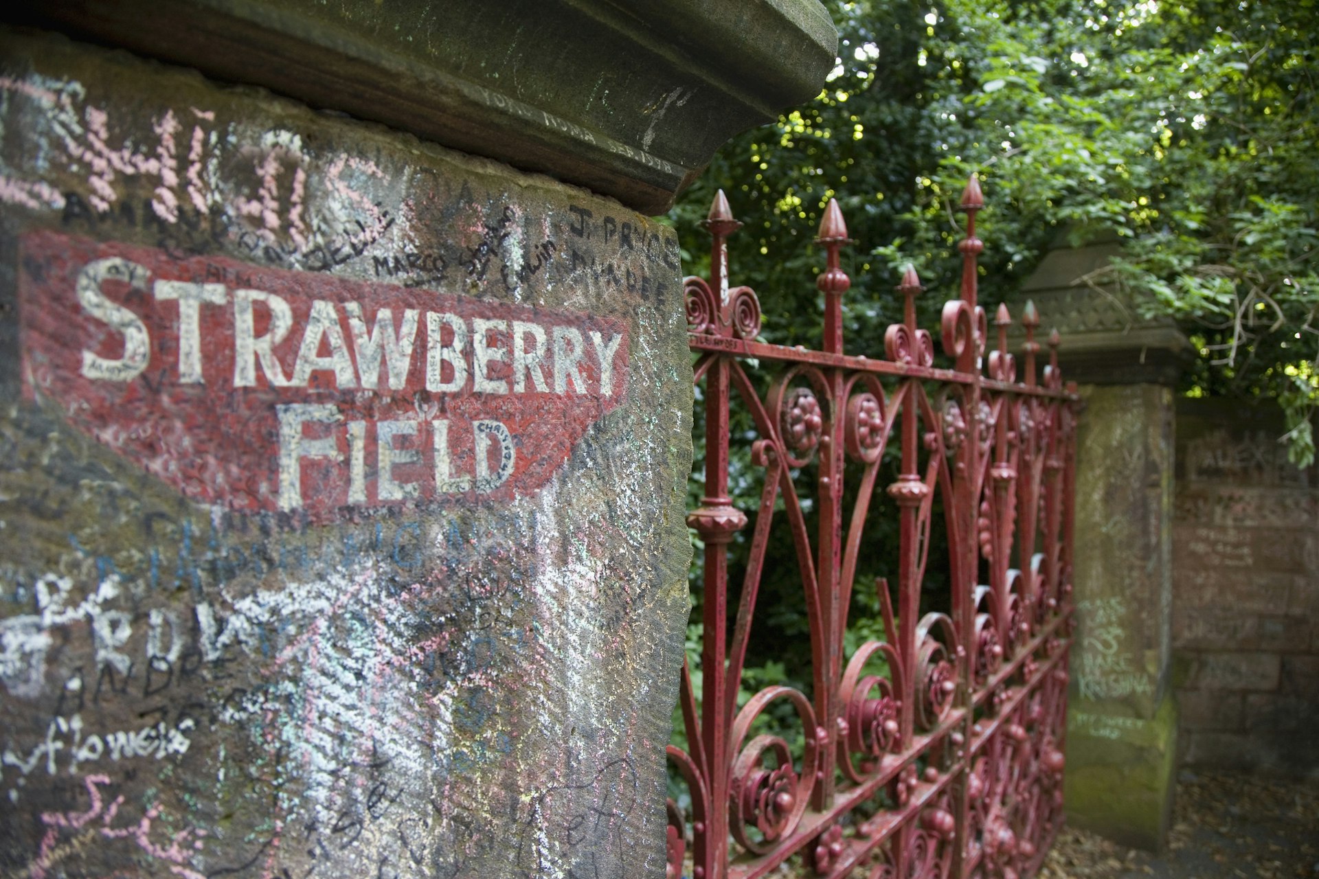 A red wrought-iron gate with graffiti on the adjacent wall - Strawberry Field gate