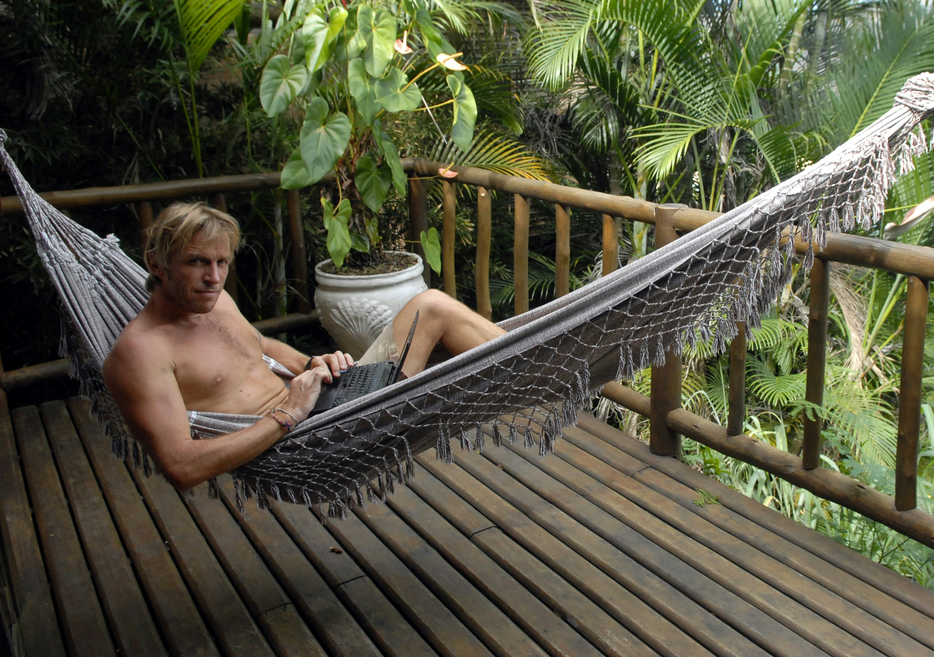 A man lies in a hammock on a wooden deck with his laptop
