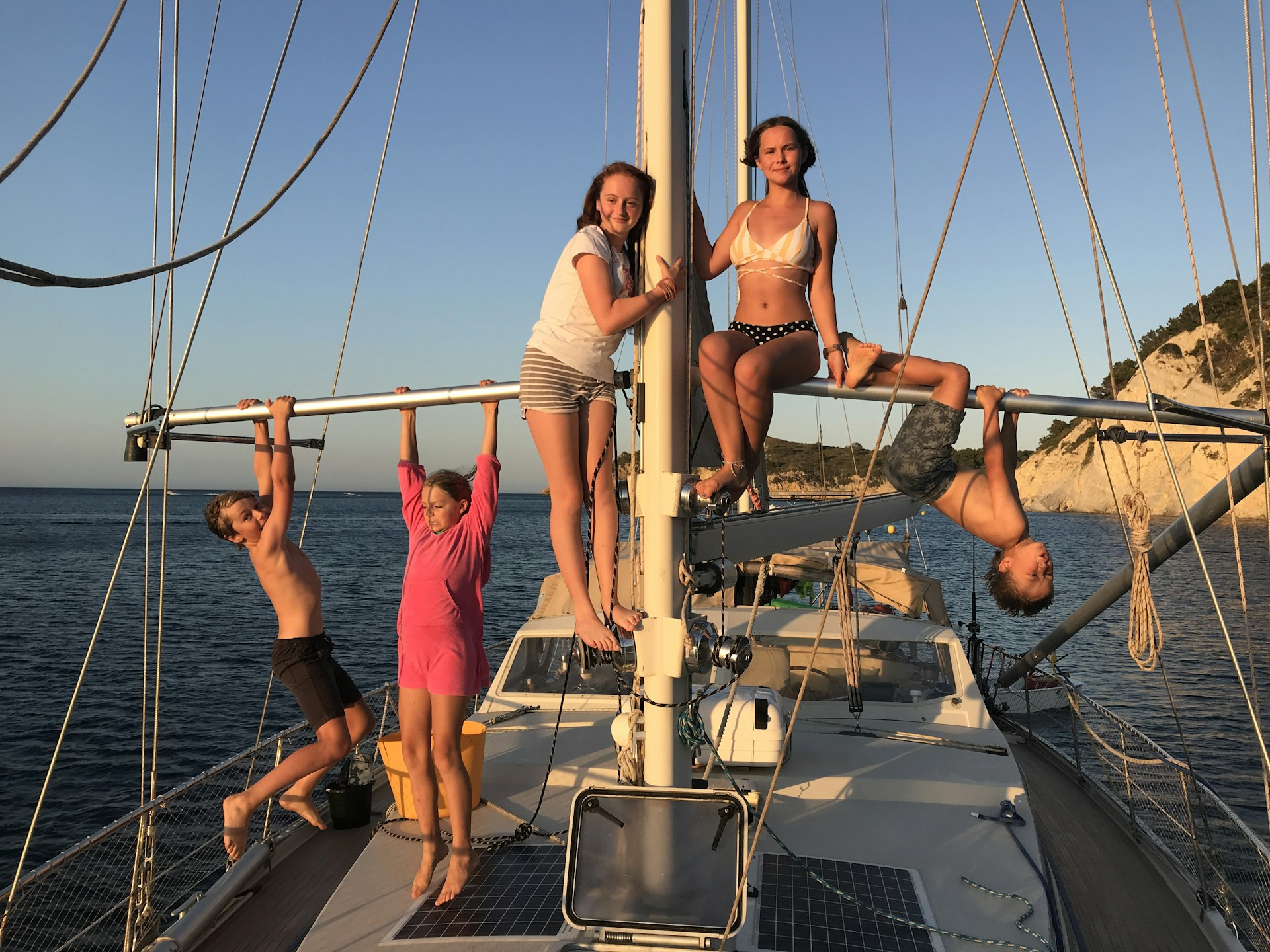 Three kids hang from the main mast on a yacht, with two other people sitting on it in the centre
