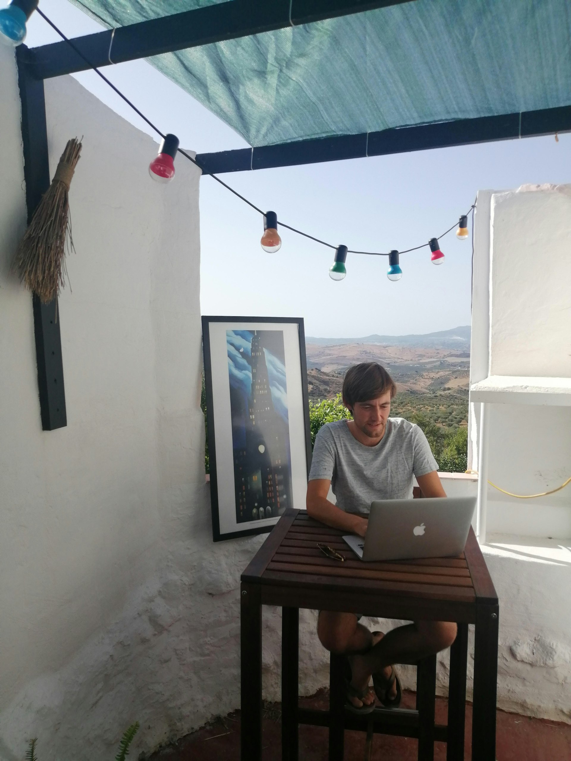 A man sits at a desk with a laptop. Behind him is a stunning view of olive groves. A painting by artist Georgia O'Keeffe is propped against the wall