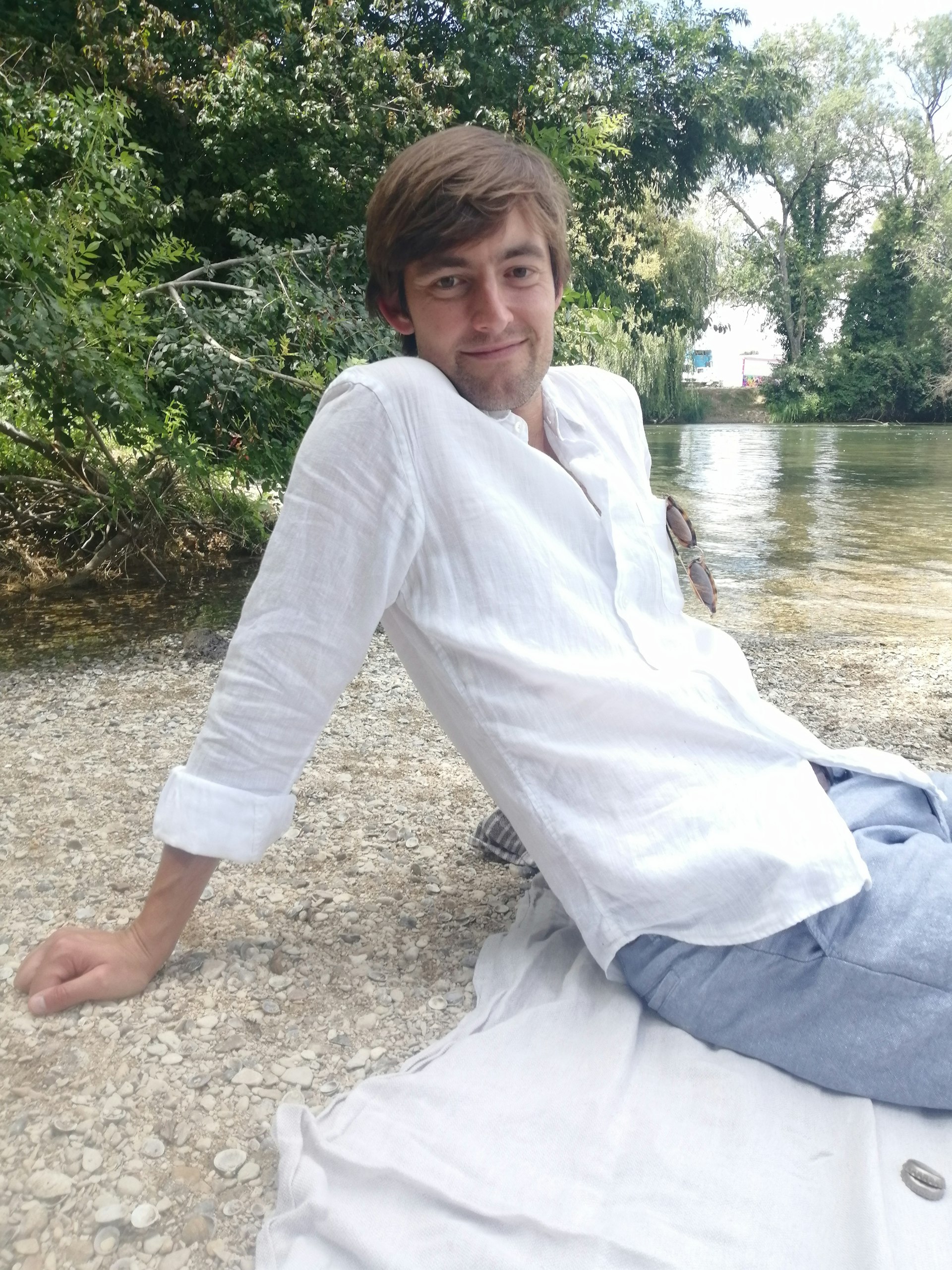 A white man with brown hair sits by a river smiling at the camera