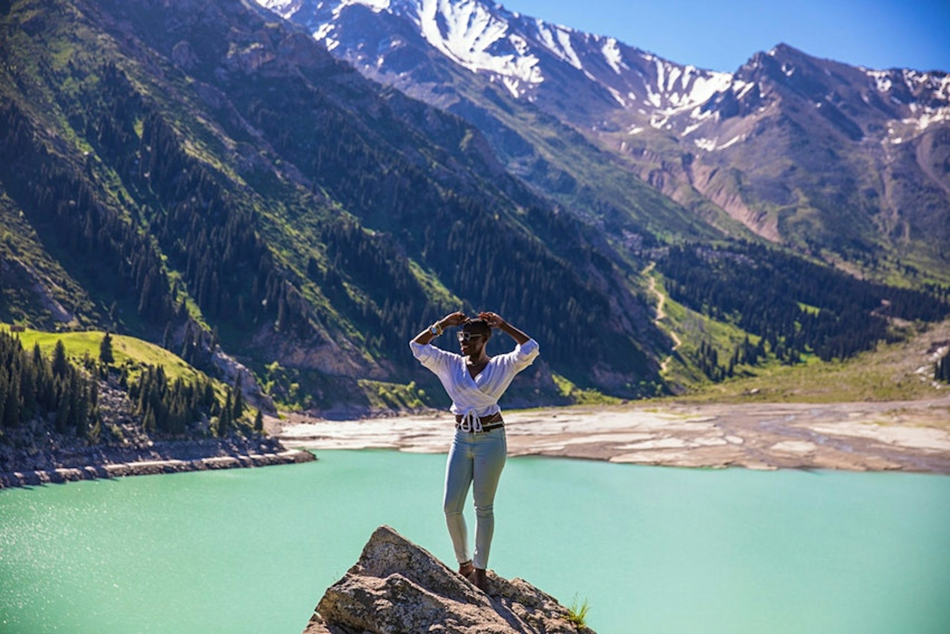 A black woman stands on a large rock above a stunning clear blue lake in a mountain landscape
