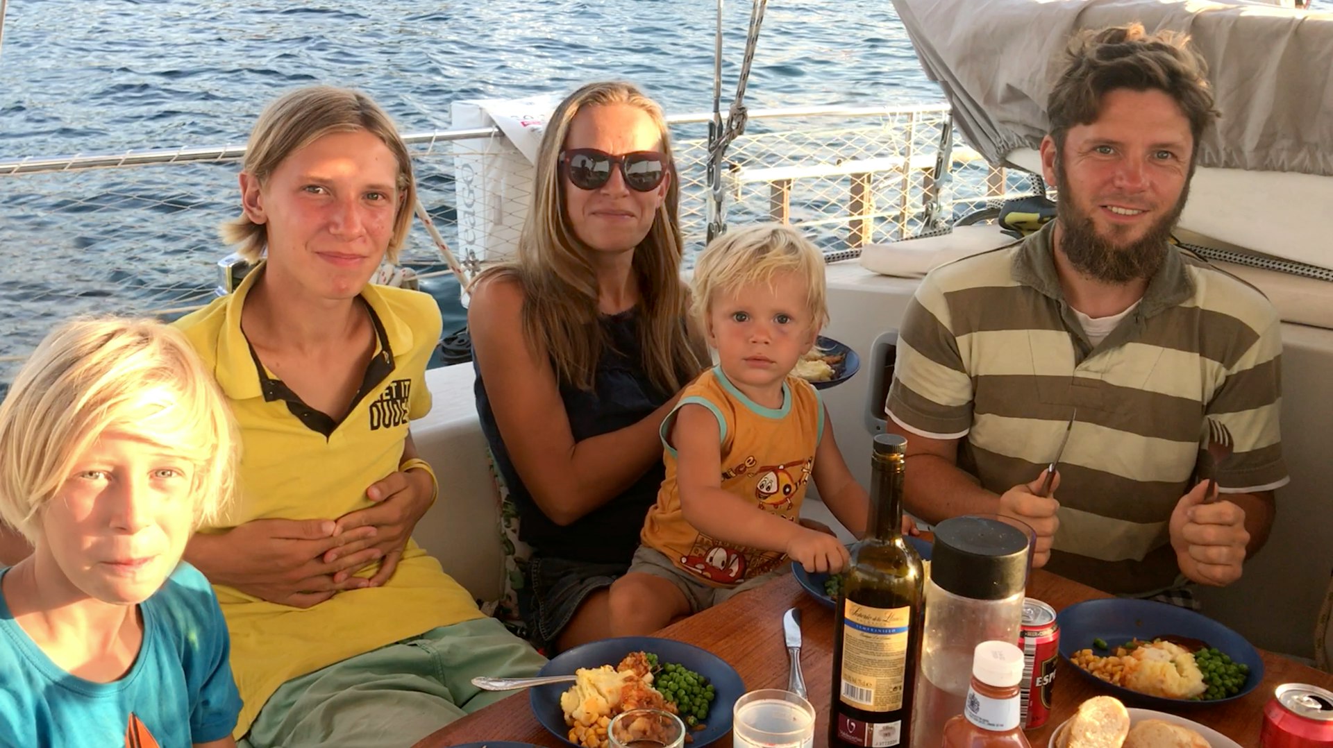 A family of two adults and three children sit eating a meal on the deck of their sailing yacht