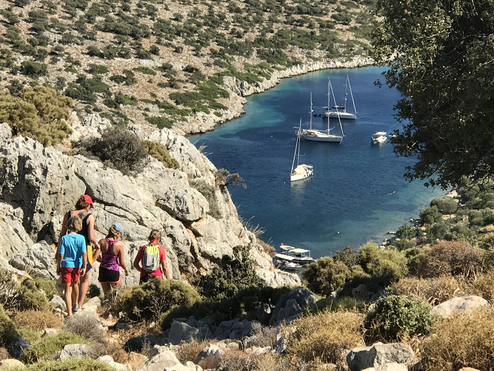 Four boats are anchored in a bay. A family walks down a rocky path towards their boat.