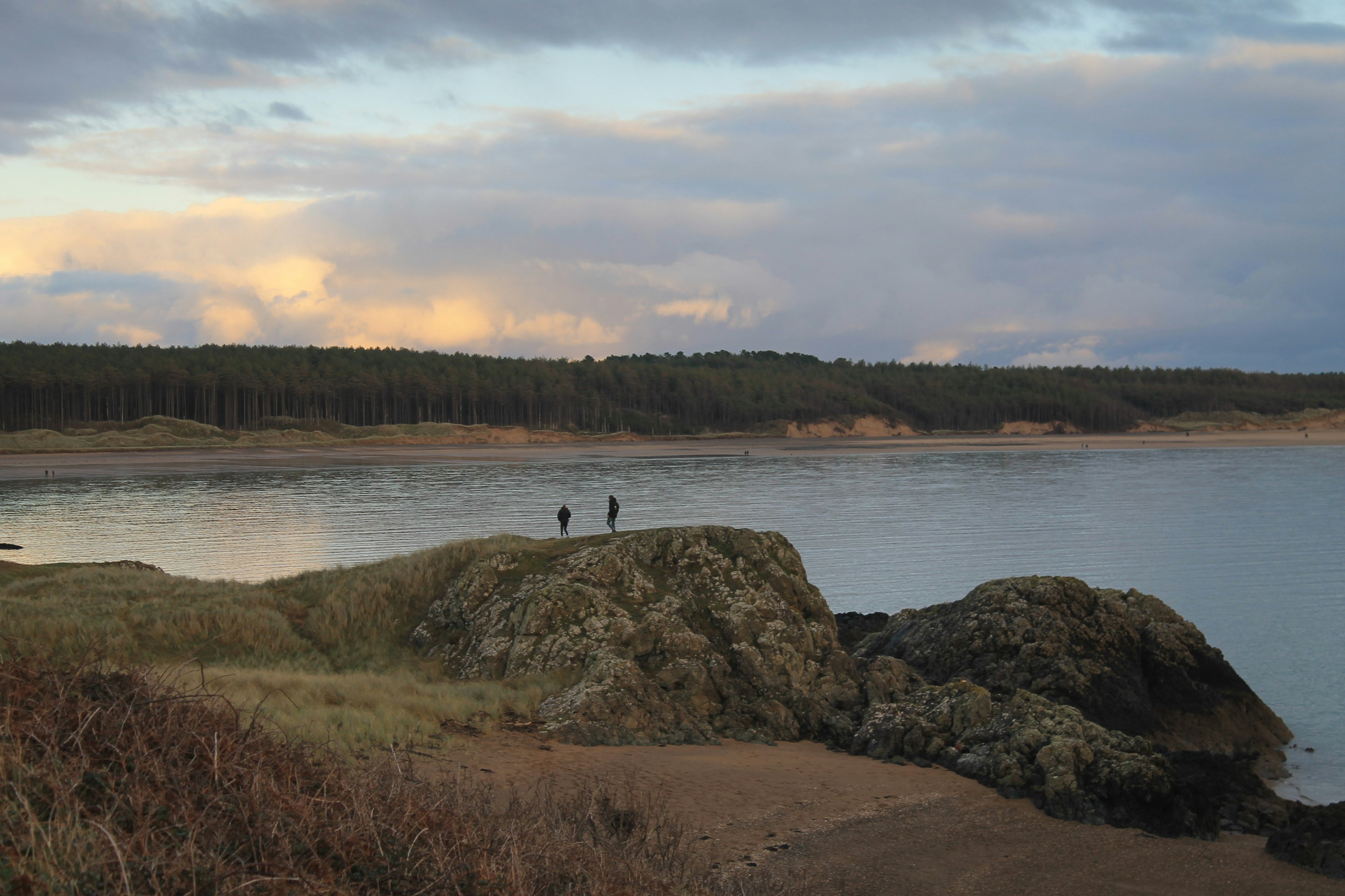 Two people walk on a hilly section of Llanddwyn Island with Newborough Beach in the background.