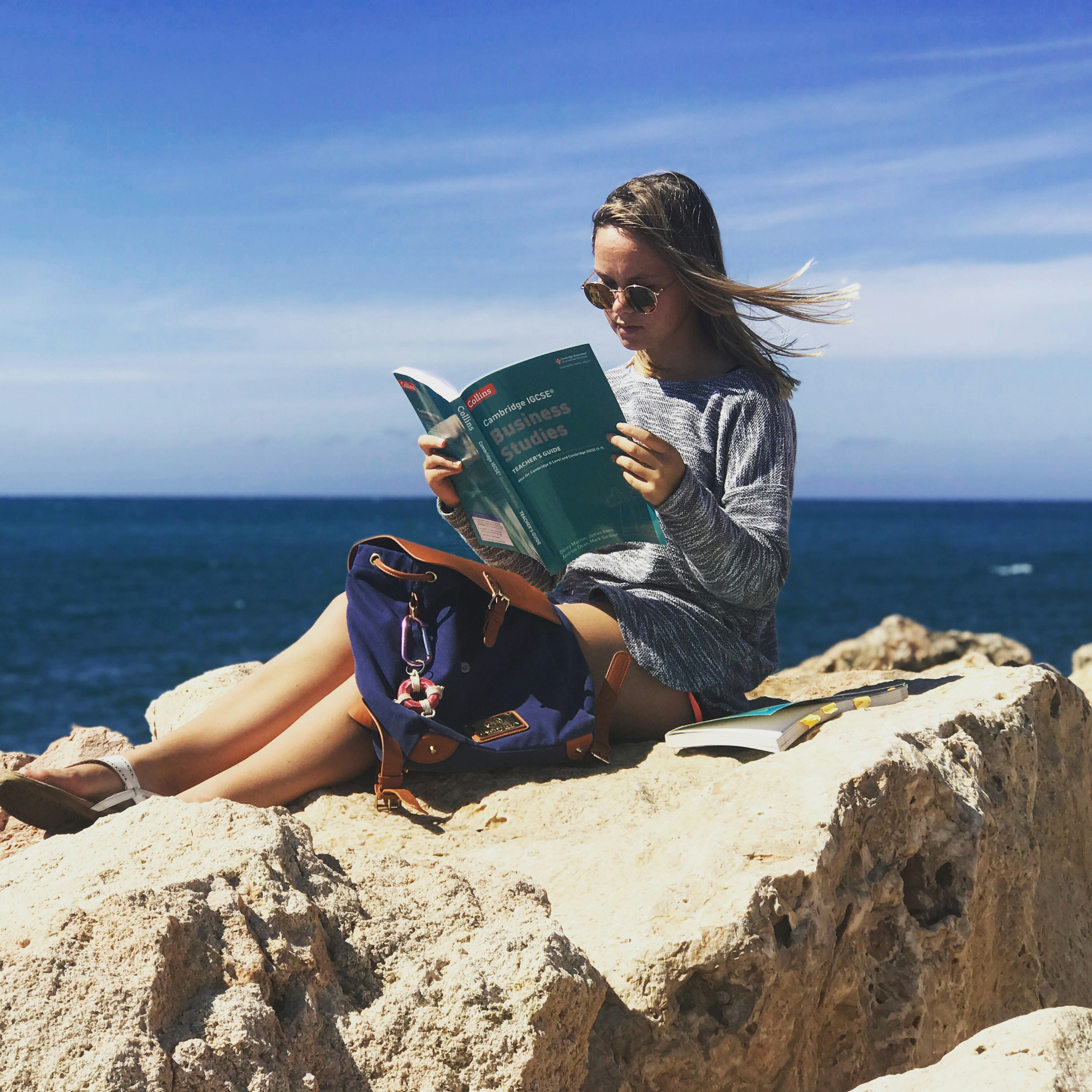 A teenage girl sits on a rock with a backpack at her feet. She's reading a textbook with the title IGCSE Cambridge Business Studies