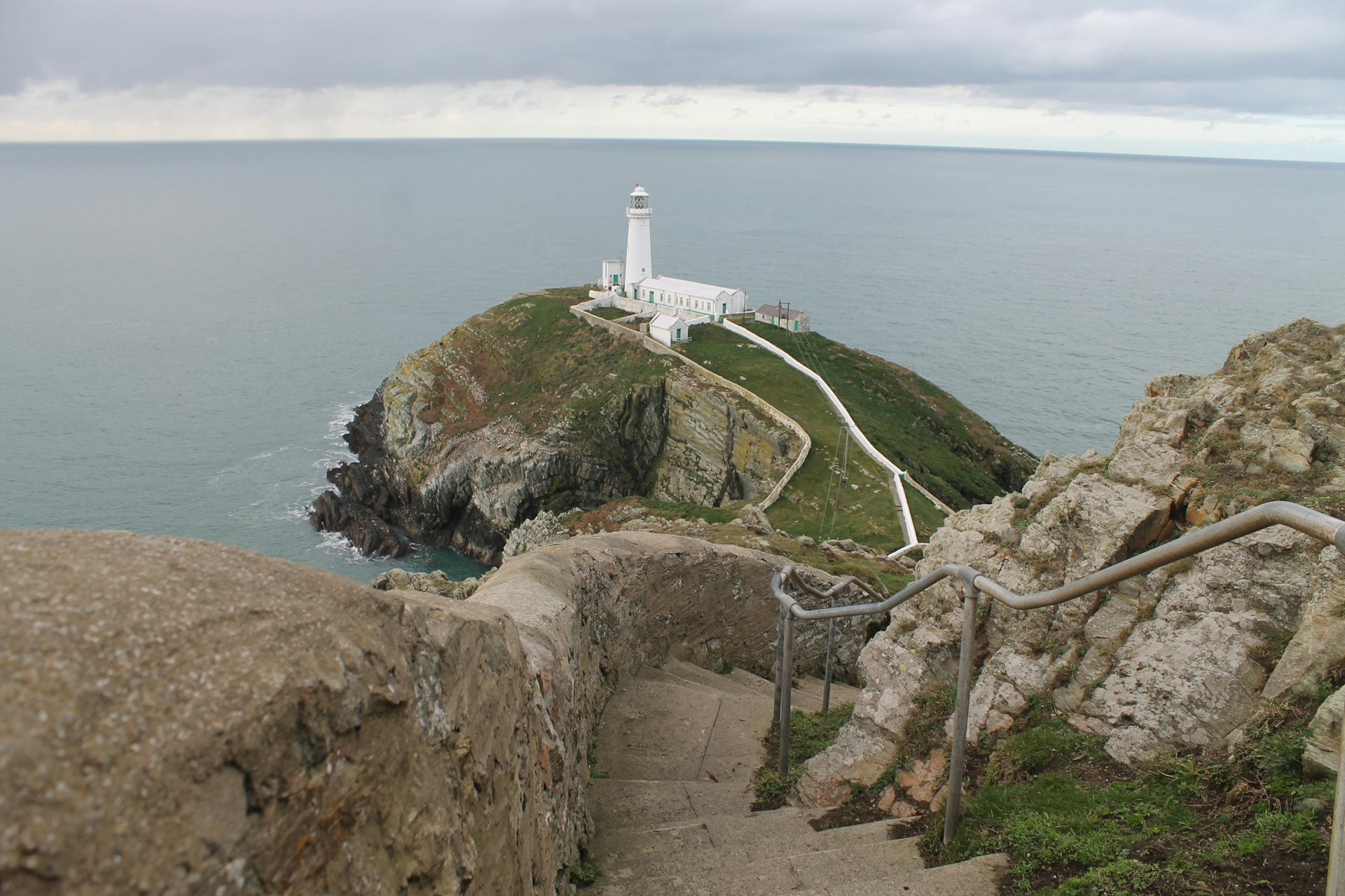 South Stack Lighthouse stands on a small rocky outcrop on the island of Anglesey. It is surrounded by sea and a cloudy sky.