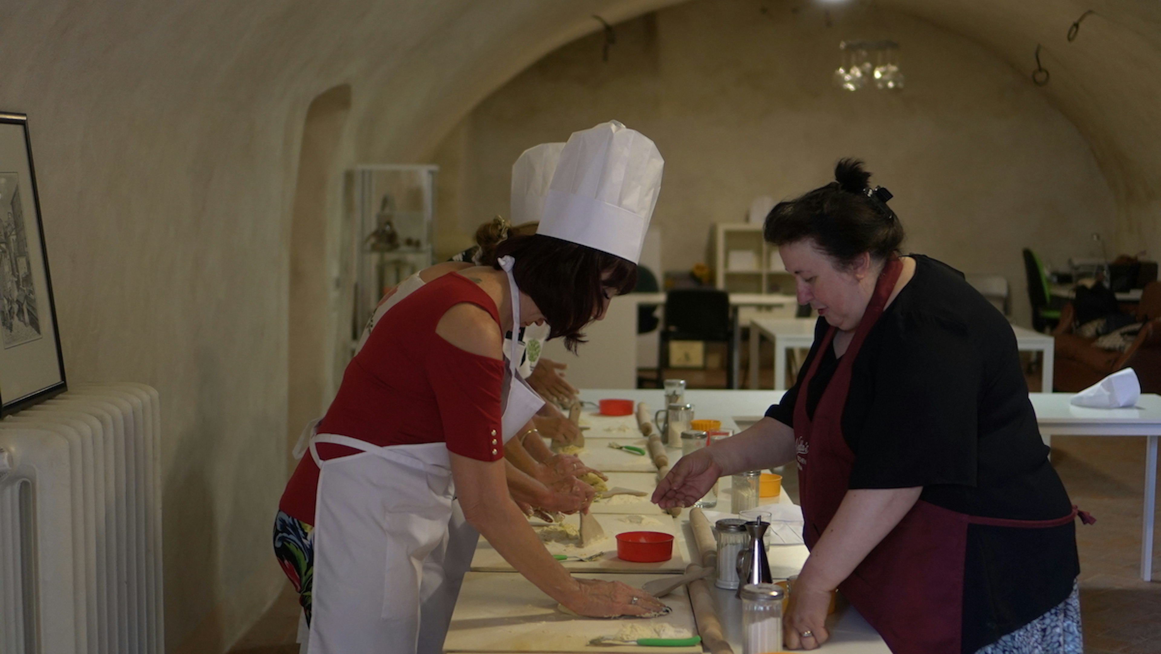 Two women, one wearing a chef's hat, lean over a counter prepping food.