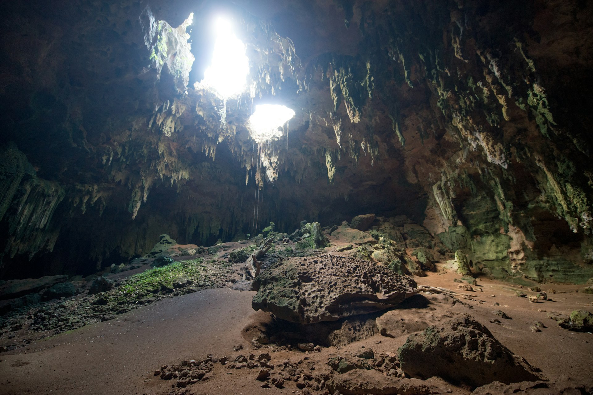 A dark cave with stalactites hanging down, and two large holes in the cave roof allowing some light to come in