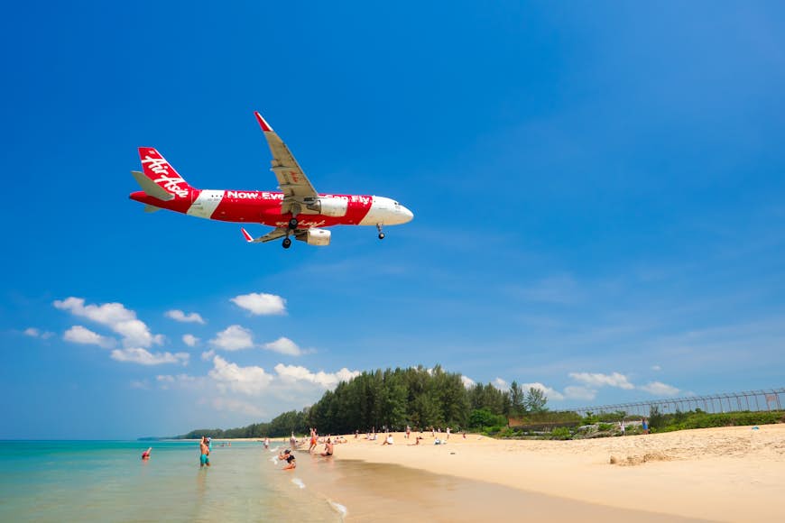 A red-and-white plane flies low over a golden sand beach on approach to the runway