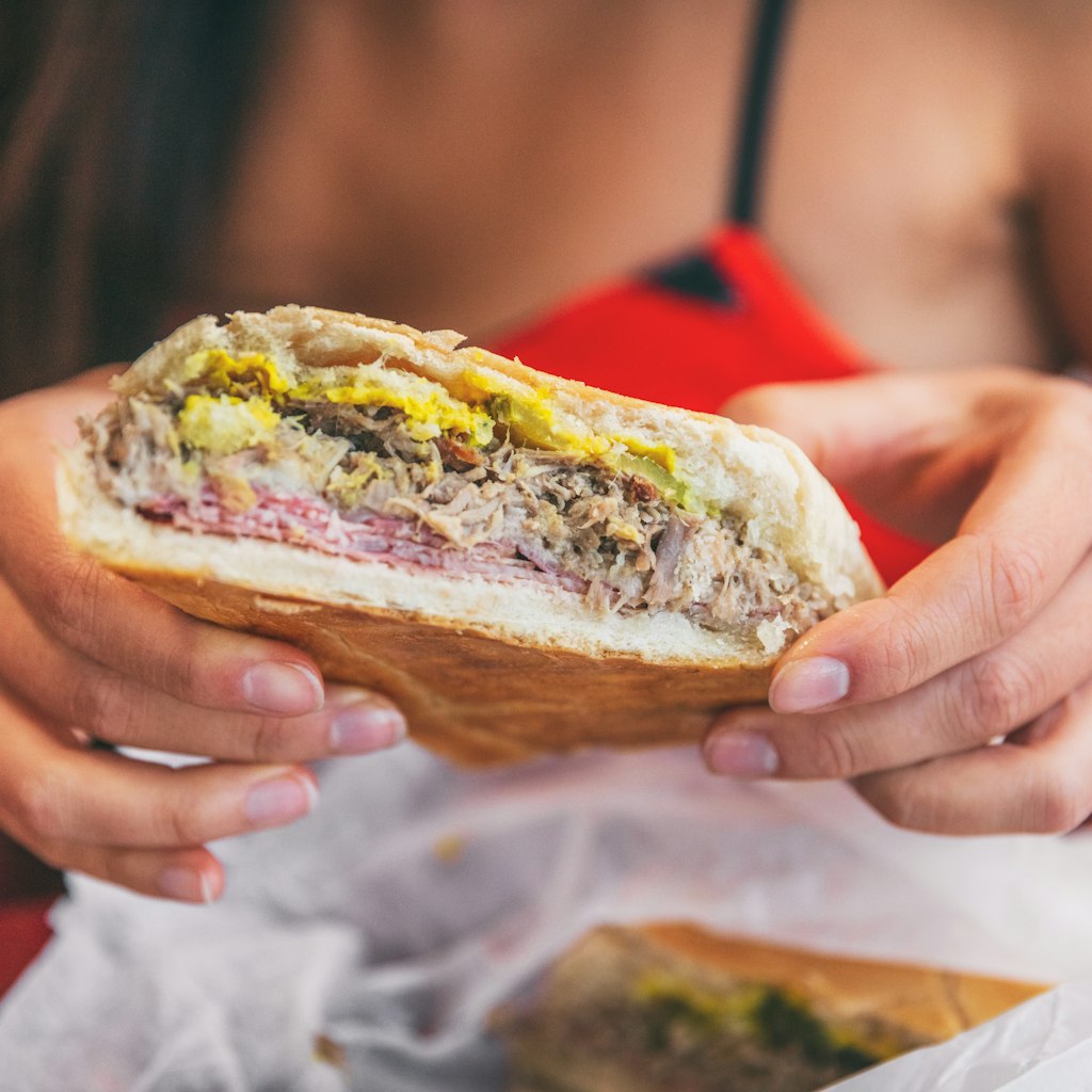 Close-up of a woman holding a Cuban sandwich outside a cafe. The sandwich consists of pressed cuban bread with roasted pork, salami sausage, swiss cheese and mustard.