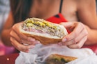 Close-up of a woman holding a Cuban sandwich outside a cafe. The sandwich consists of pressed cuban bread with roasted pork, salami sausage, swiss cheese and mustard.