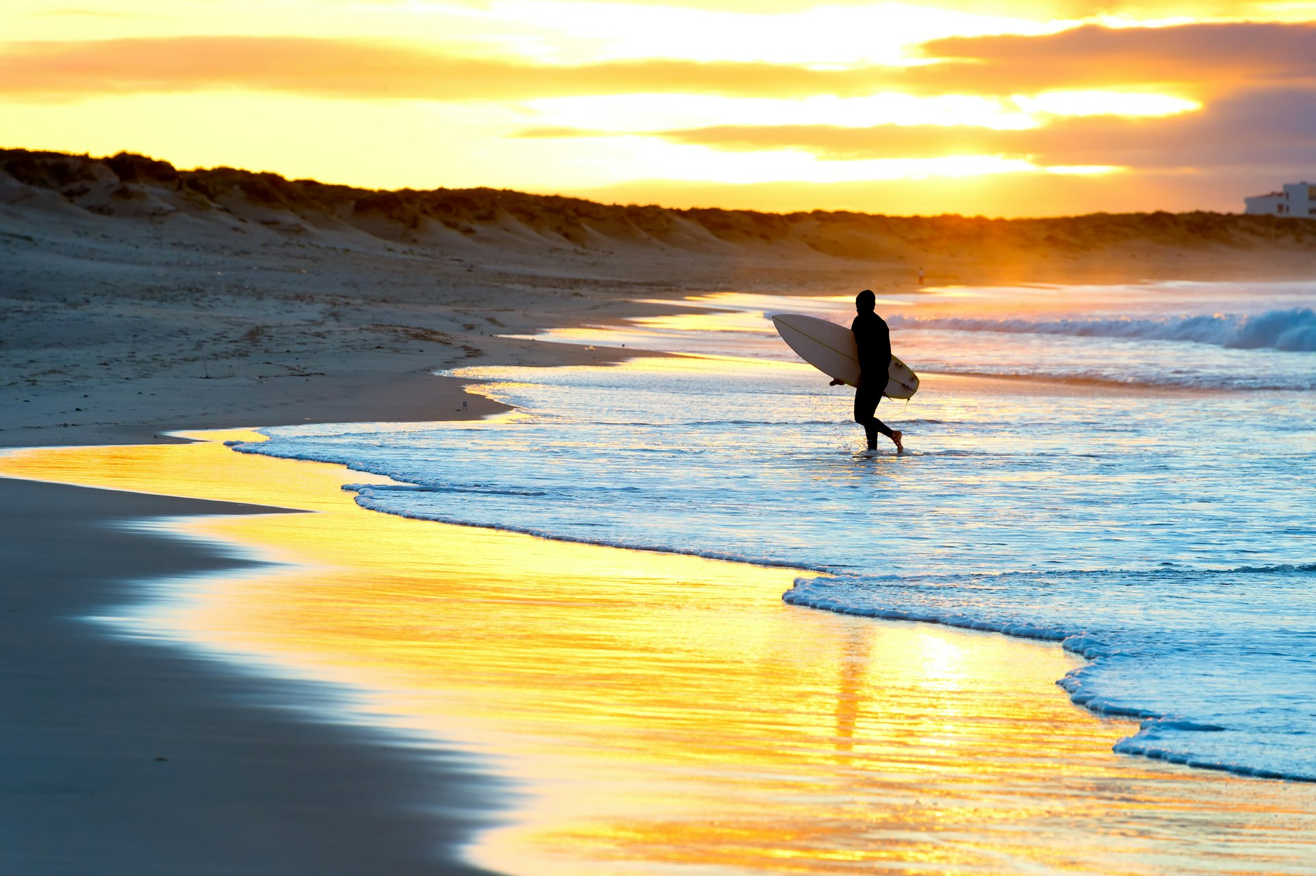 A surfer carrying a surfboard on the beach is is silhouetted against the setting sun