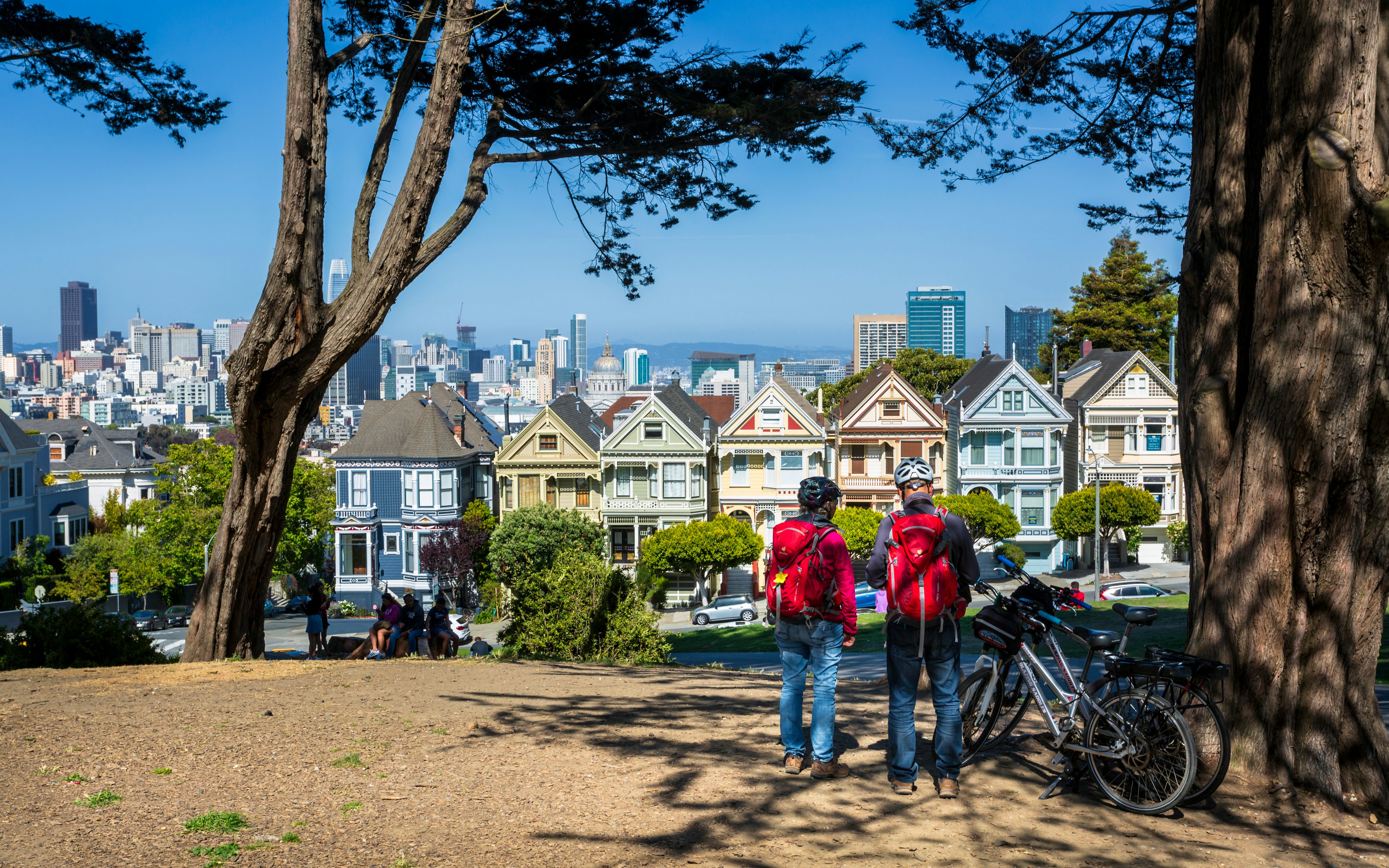 Two cyclists stand next to their bikes looking at a view of pastel-colored houses with a cityscape behind