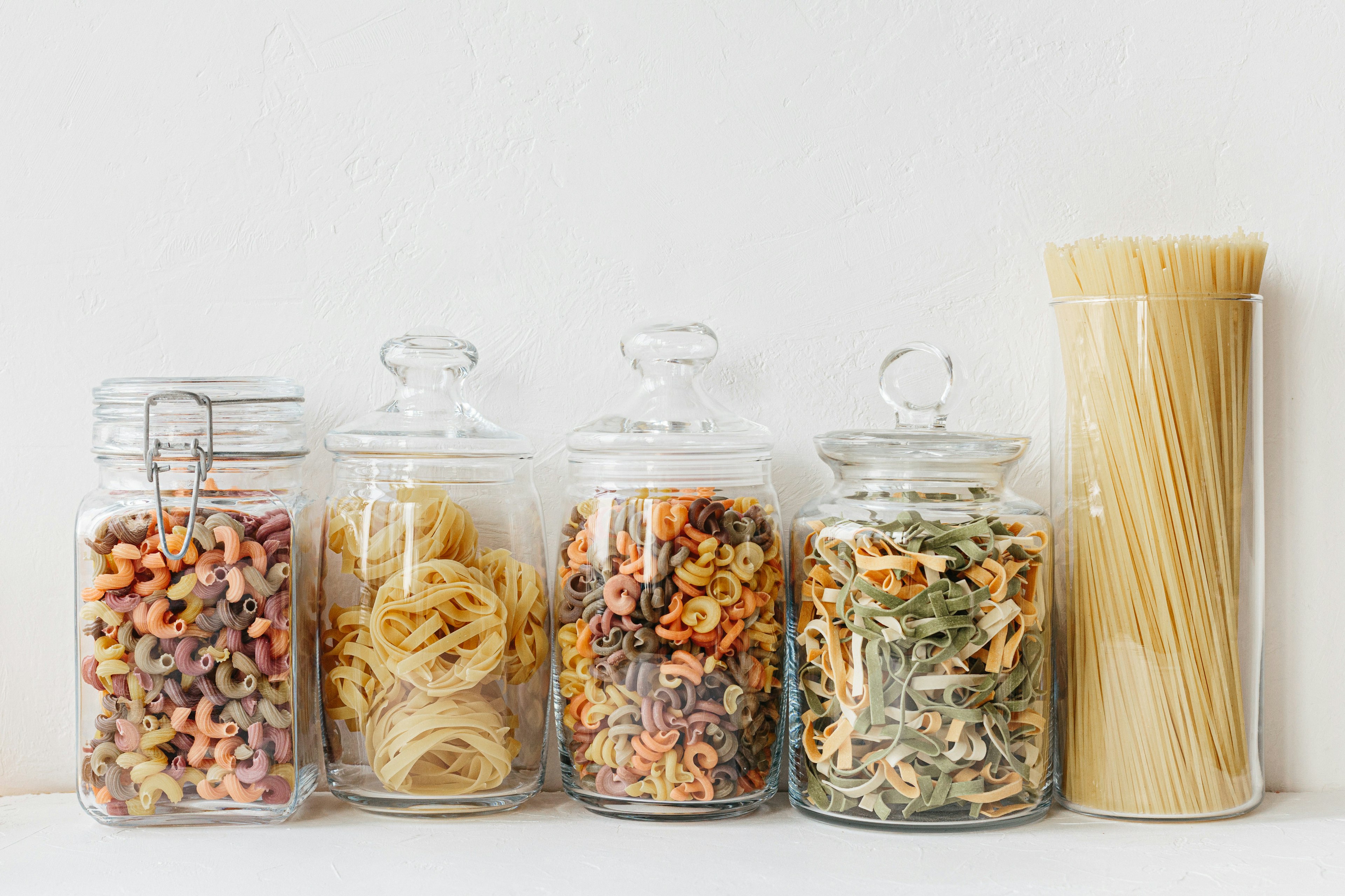 Four glass jars filled with dried pasta lined up against a white wall