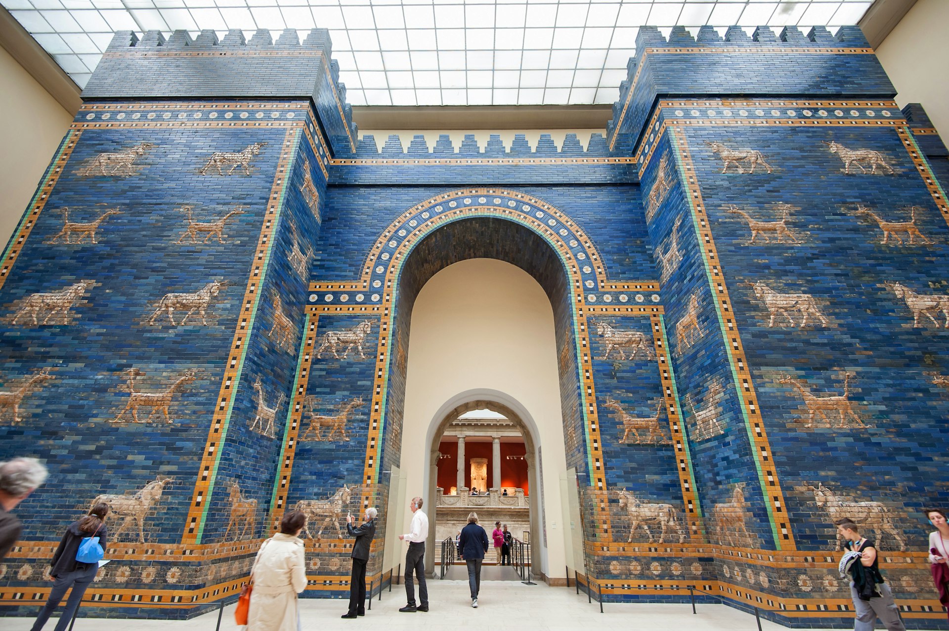 Visitors walk by or stand in front of bright blue walls of Babylonian city inside the Pergamon museum.