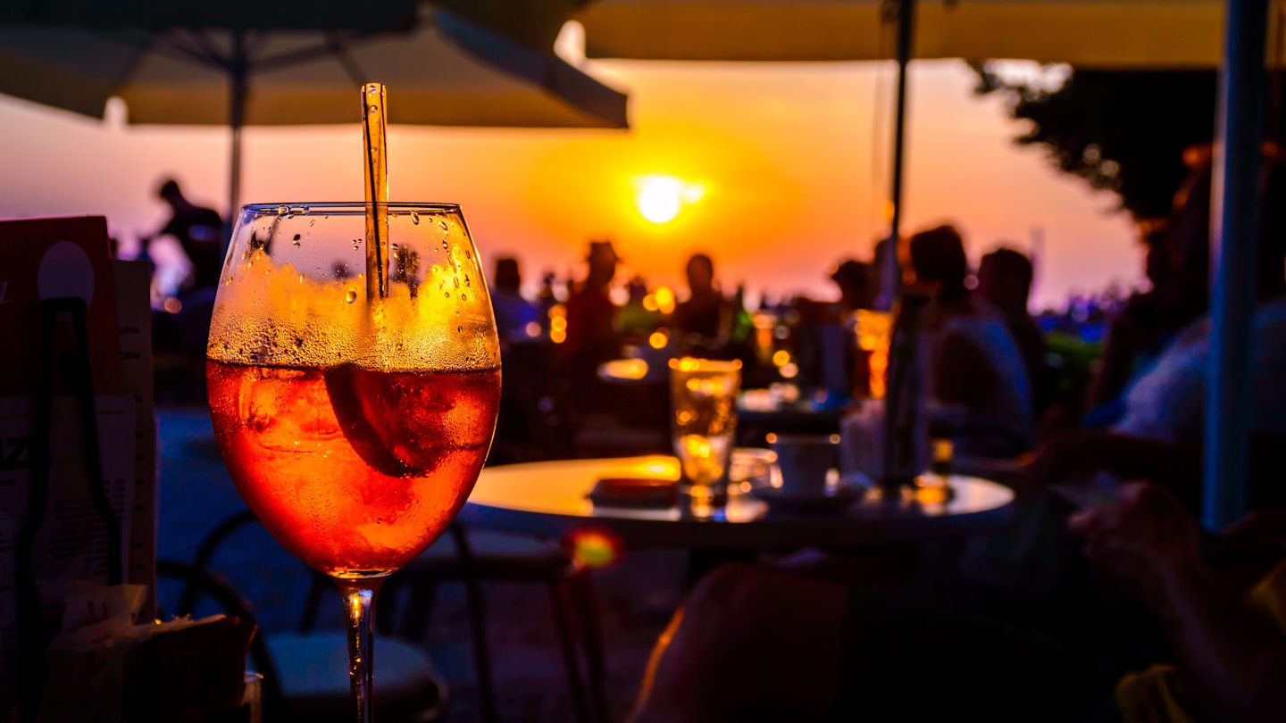 A glass of cold orange cocktail at the sunset on the table of a beach bar at the sunset, with blurry people around having refreshments or partying on a summer evening, with copy space for text