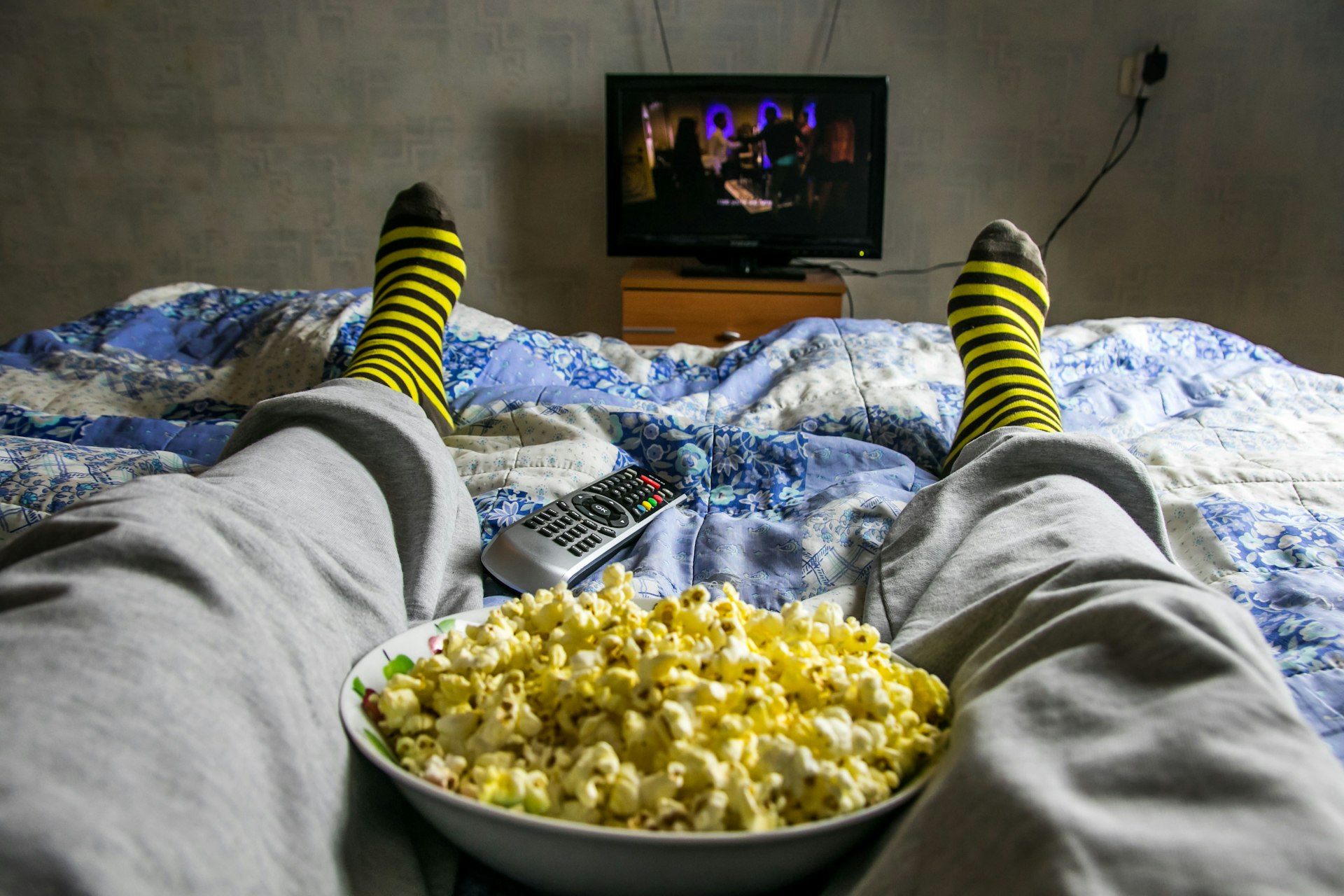 Legs of a person laying on a bed, wearing gray pants and stripe socks has a bowl of popcorn between their legs and a remote control by their foot. There is a TV on a wooden stand.
