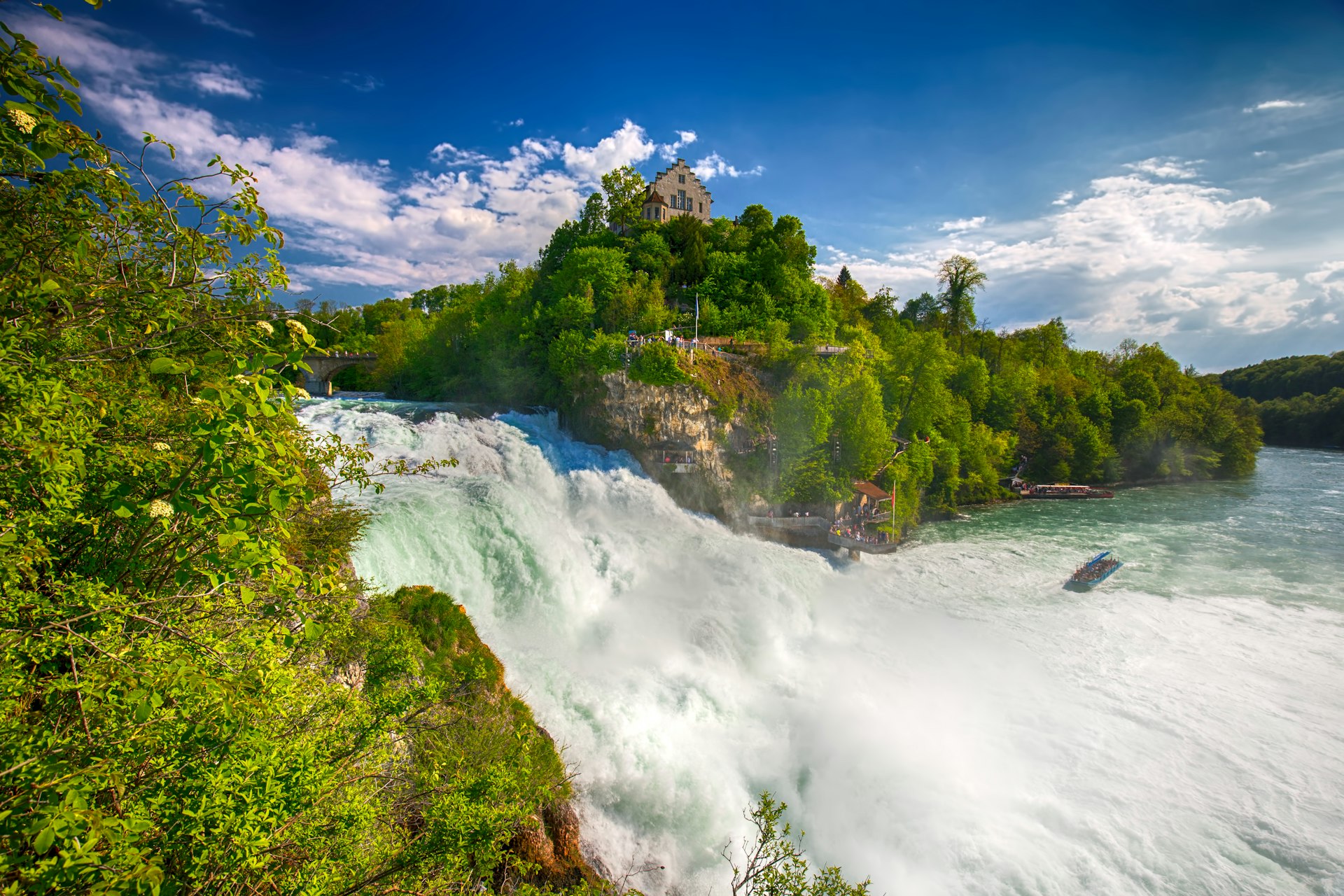 View of a vast and energetic waterfall with people standing at a viewpoint backed by a small building