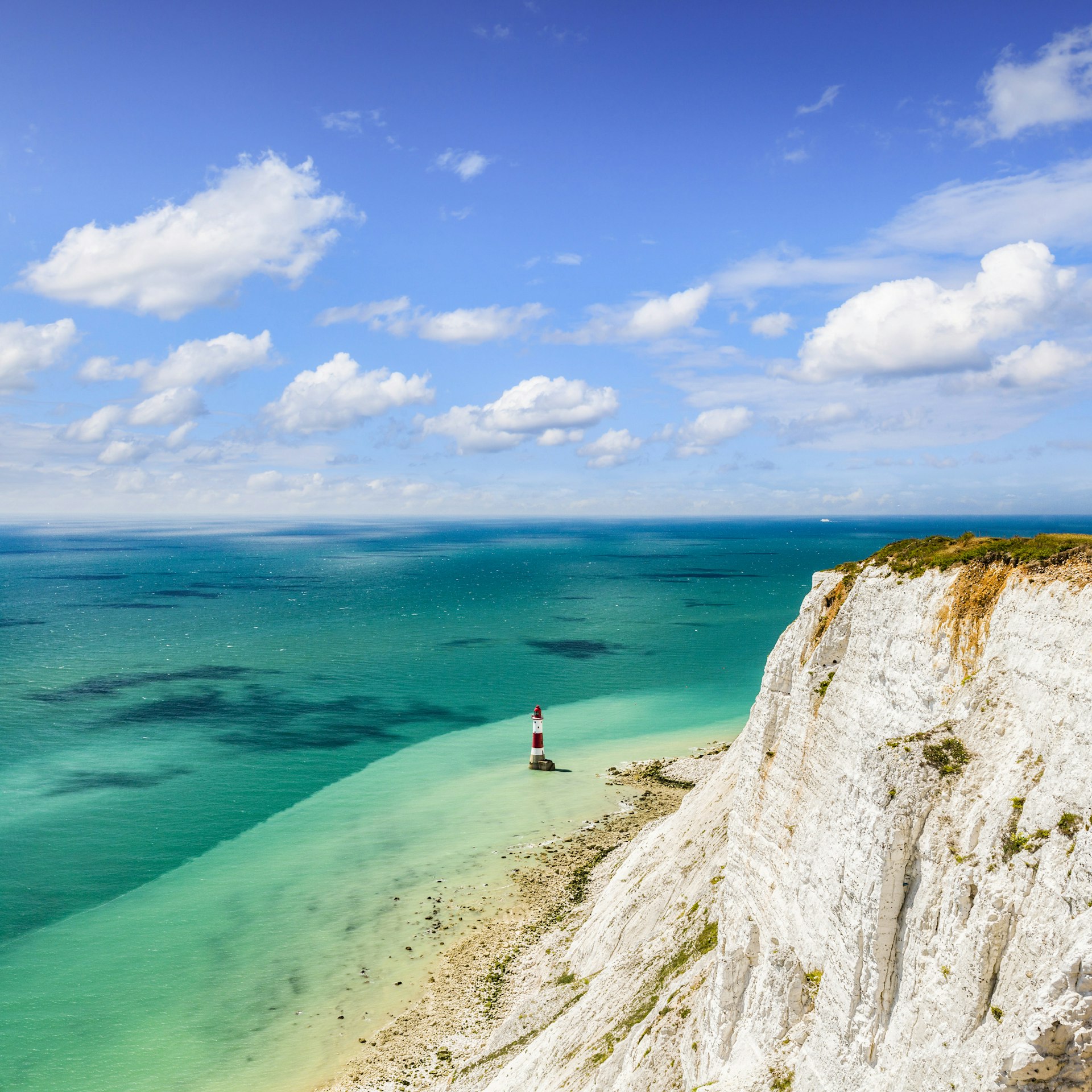 A white cliff on the edge of a turquoise sea. A red-and-white striped lighthouse stands in the shallows