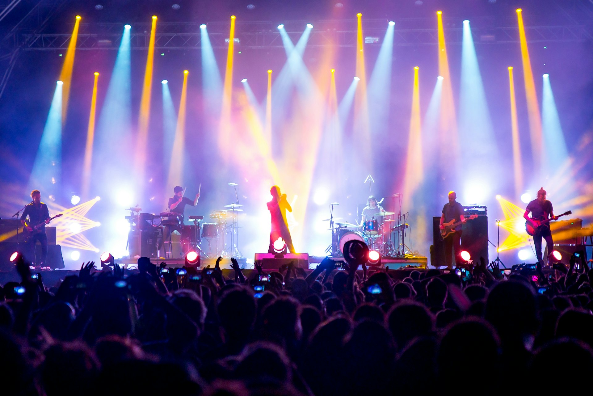 A band performing on stage is silhouetted among bright spotlights and smoke effects