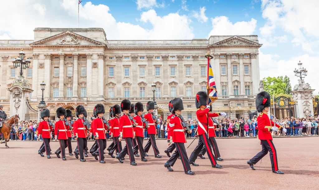 A row of soldiers in red uniforms with tall black hats march in front of a white/grey palace building 
