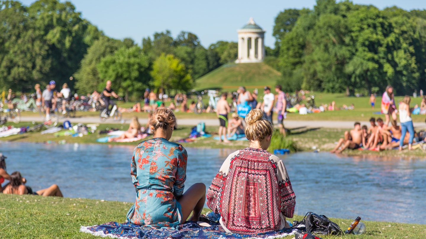 Two girls sitting on a rug at English Garden (Englischer Garten) with a crowd of others in the background.