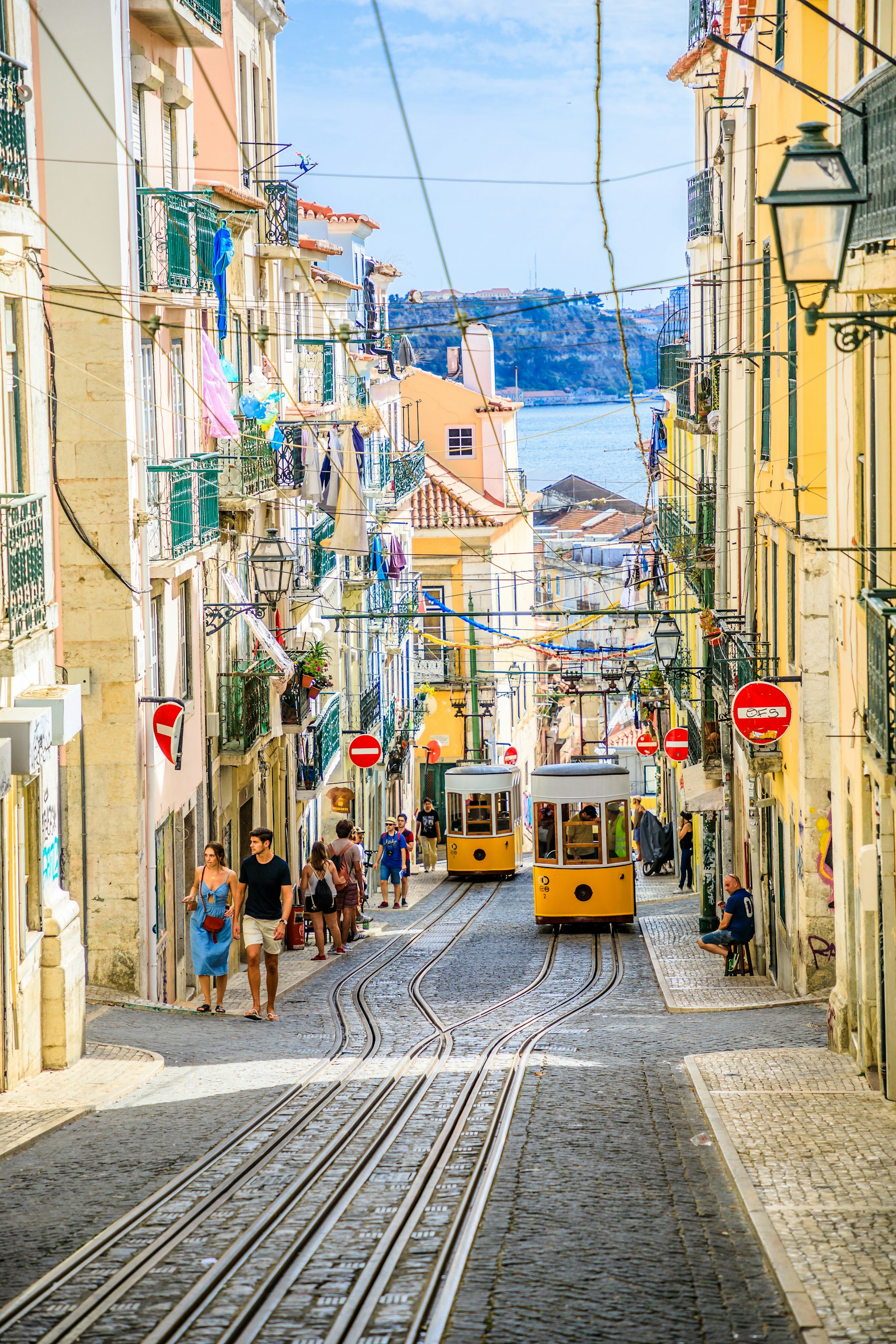 Two yellow funiculars pass each other on a hilly street with a body of water in the distance