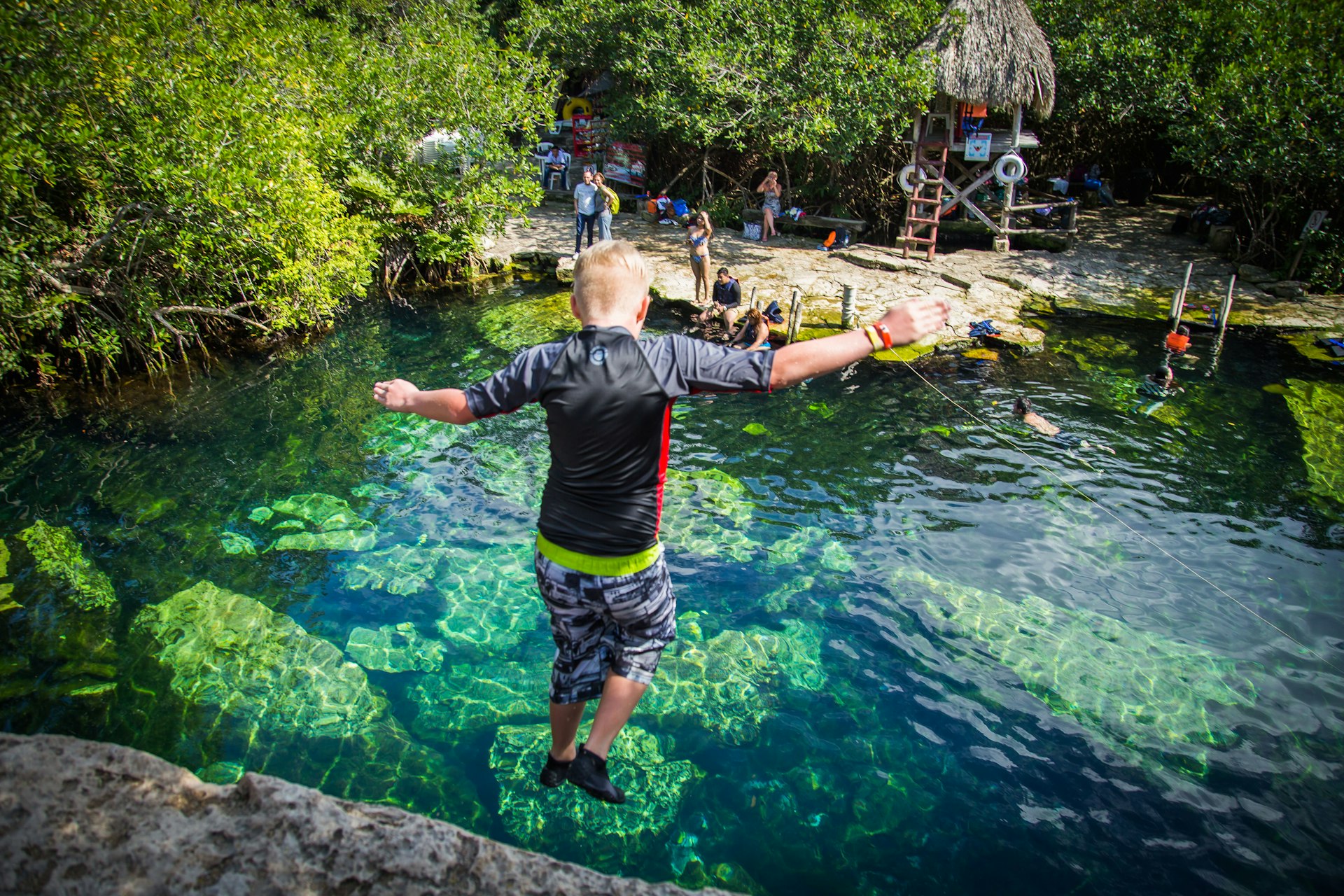A young boy jumps off a ledge into a clear blue cenote (swimming hole)