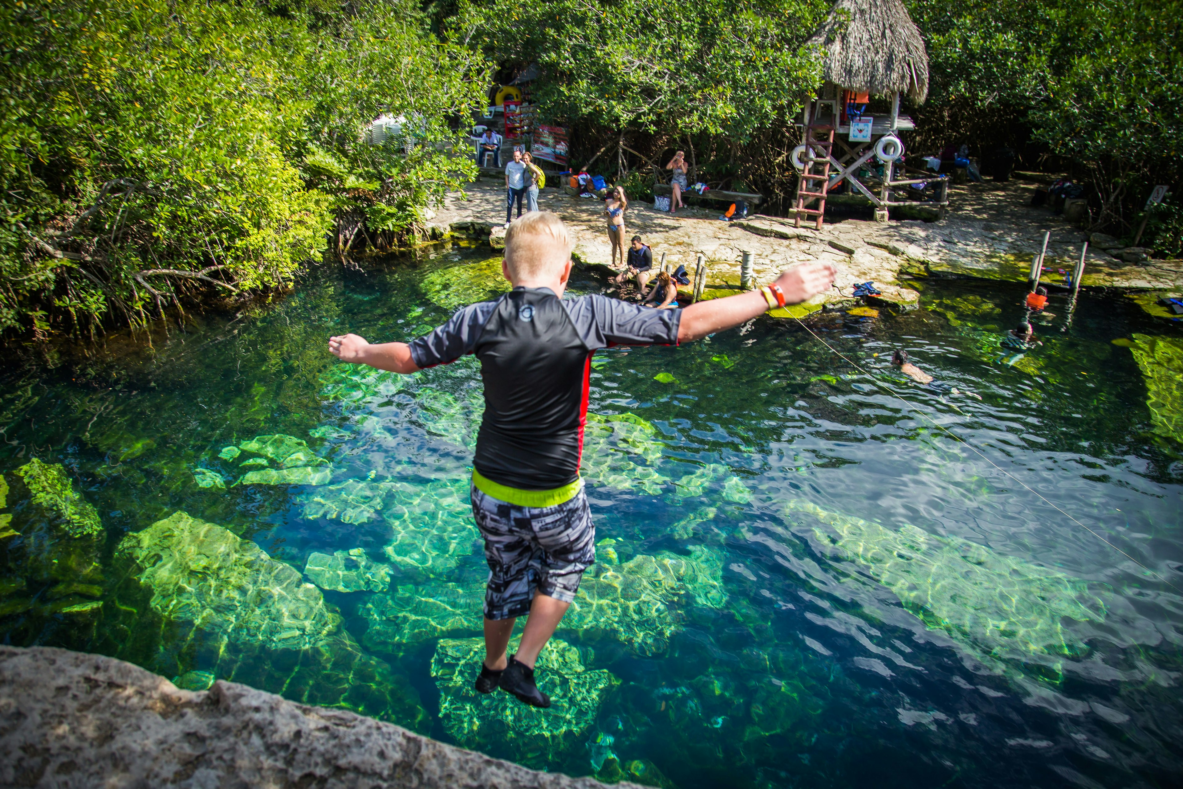 A young boy jumps off a ledge into a clear blue cenote (swimming hole)