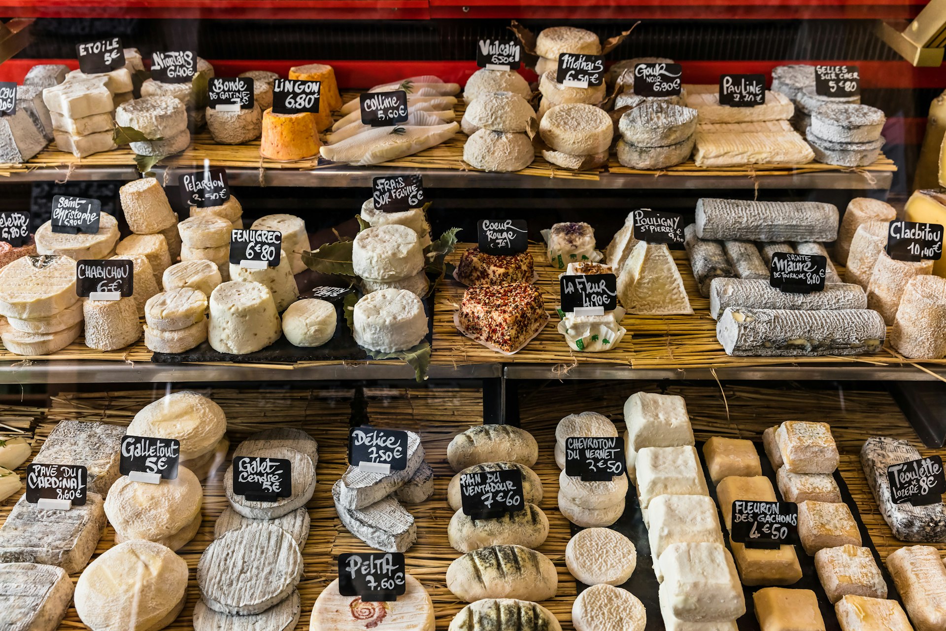 A selection of French and Italian cheeses on display in a food hall. Each one has a small blackboard near it outlining the name and price.