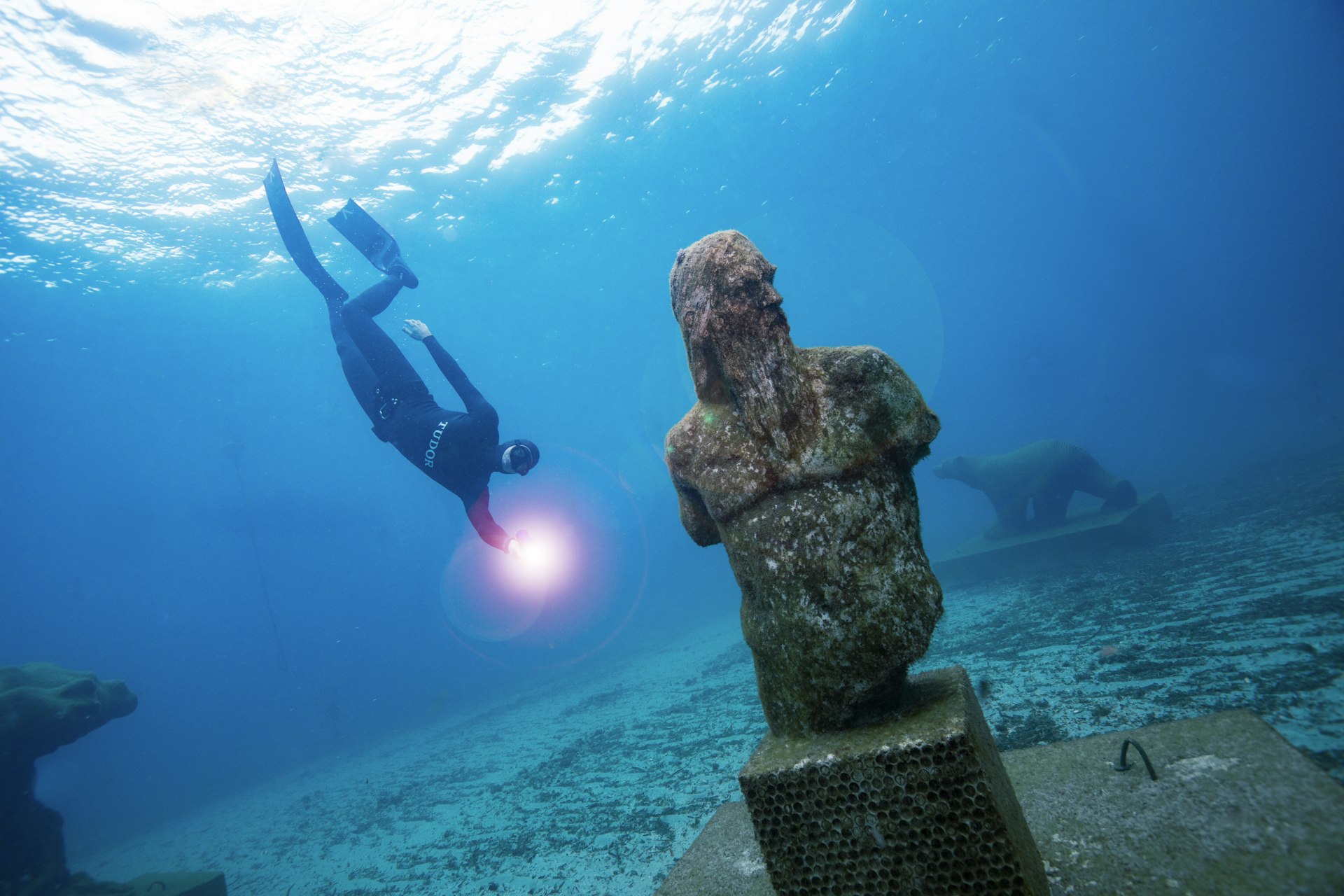 An underwater shot of a diver approaching a statue of Poseidon which stands on the seabed