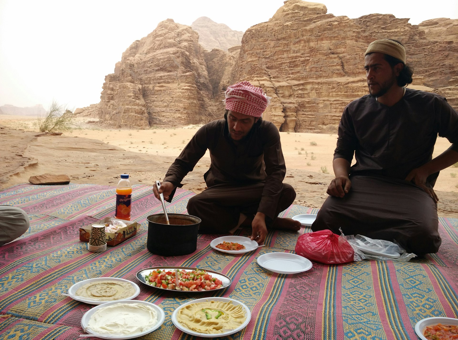 Two Bedouin men eat food on a brightly striped carpets in the desert of Jordan