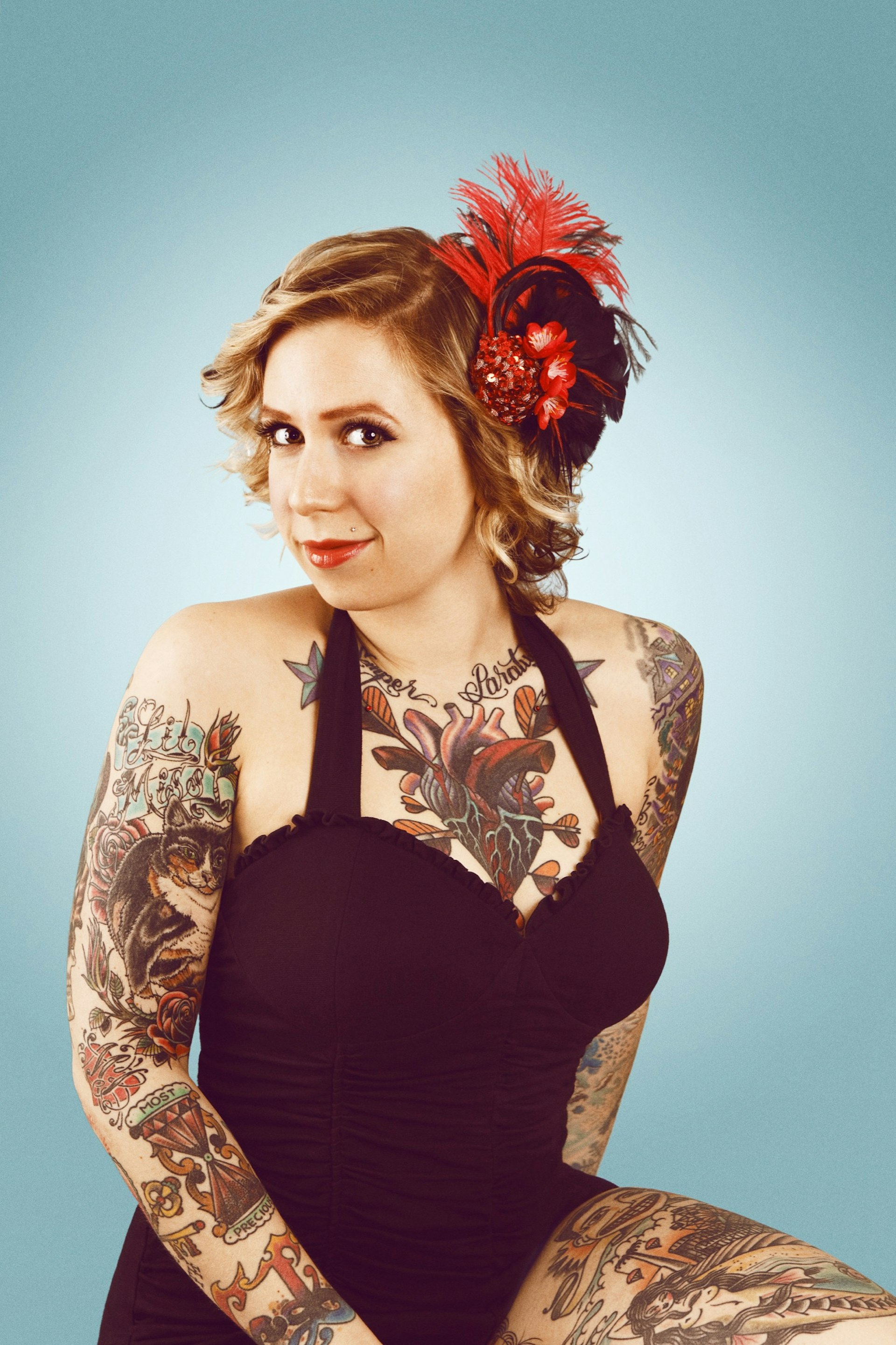 A woman wearing a black halter bathing suit is covered in American-style, colorful tattoos. She also has a large red head piece on the side of her head.