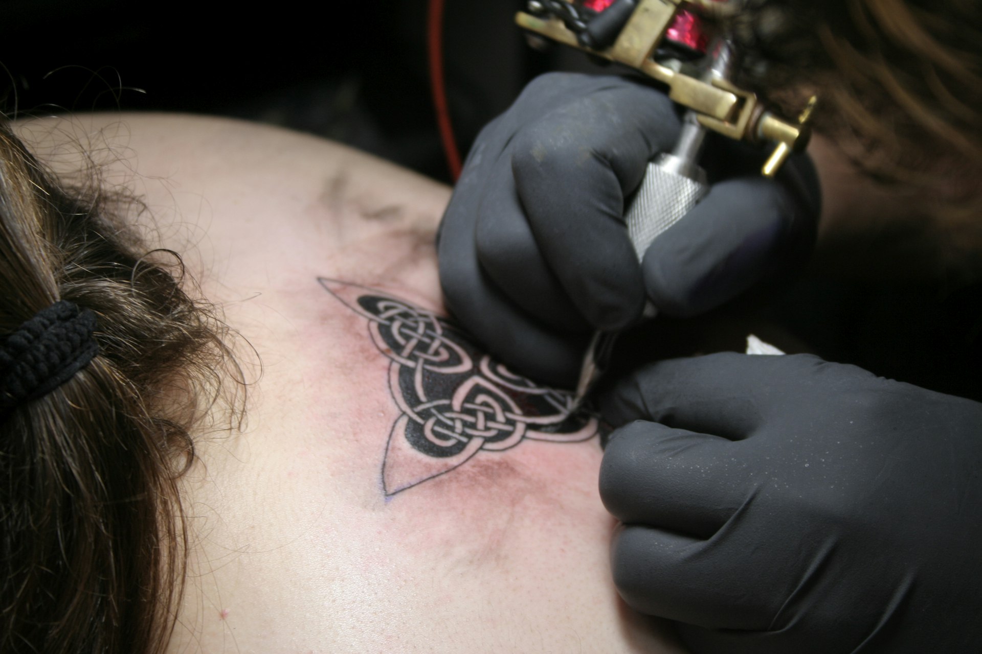 Closeup of a tattoo artist wearing black gloves and holding a tattoo machine as they ink a Celtic symbol on the back of a person