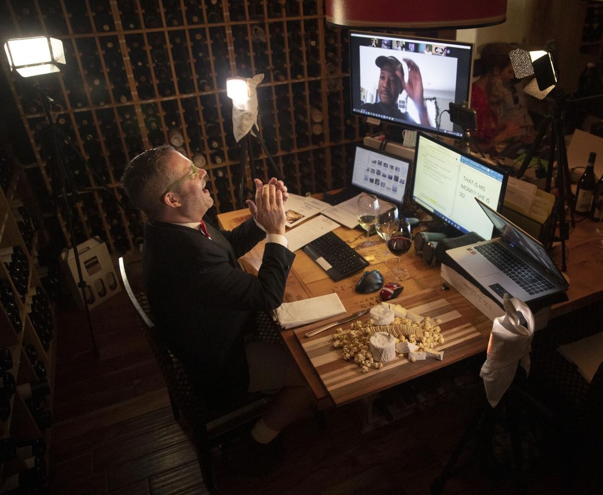 A man at a desk in front of multiple screens, laughing and eating wine and cheese