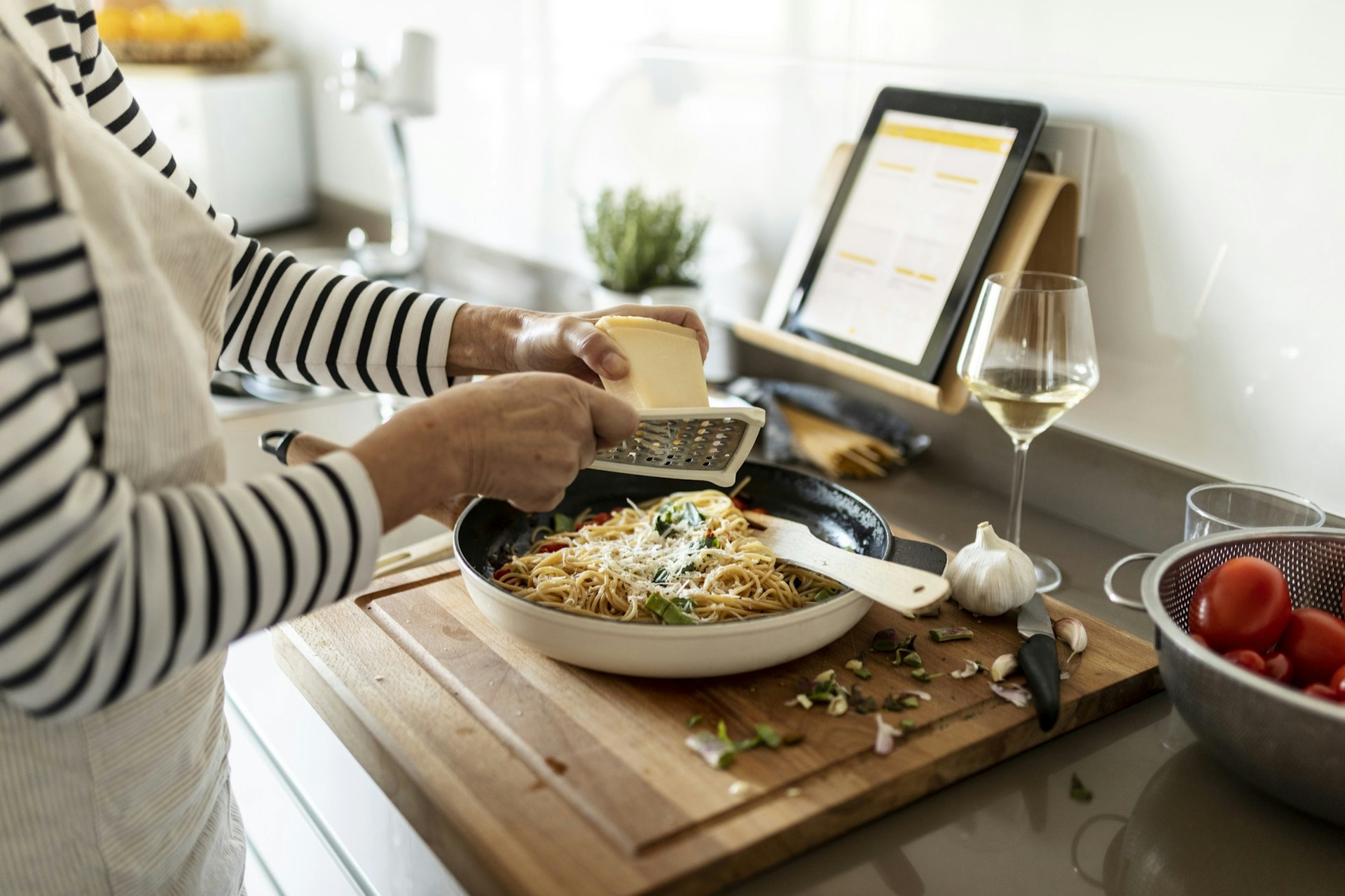 Close-up of woman with tablet shaving cheese over a pasta dish. There is a glass of wine night to the cutting board.