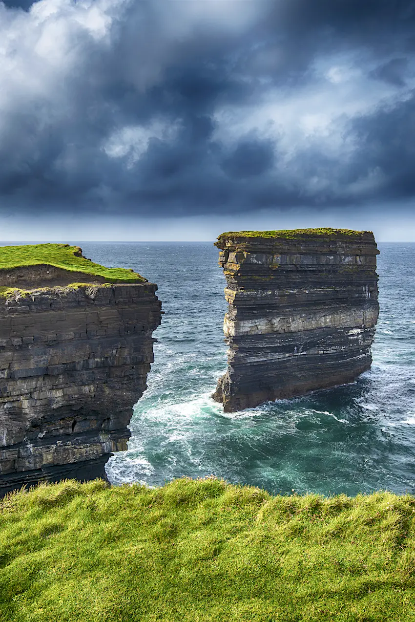 Dun Briste Downpatrick Head Waves crash at the base of the Dun Briste. There's bright green grass at the top of the natural formation on a dark cloudy day.