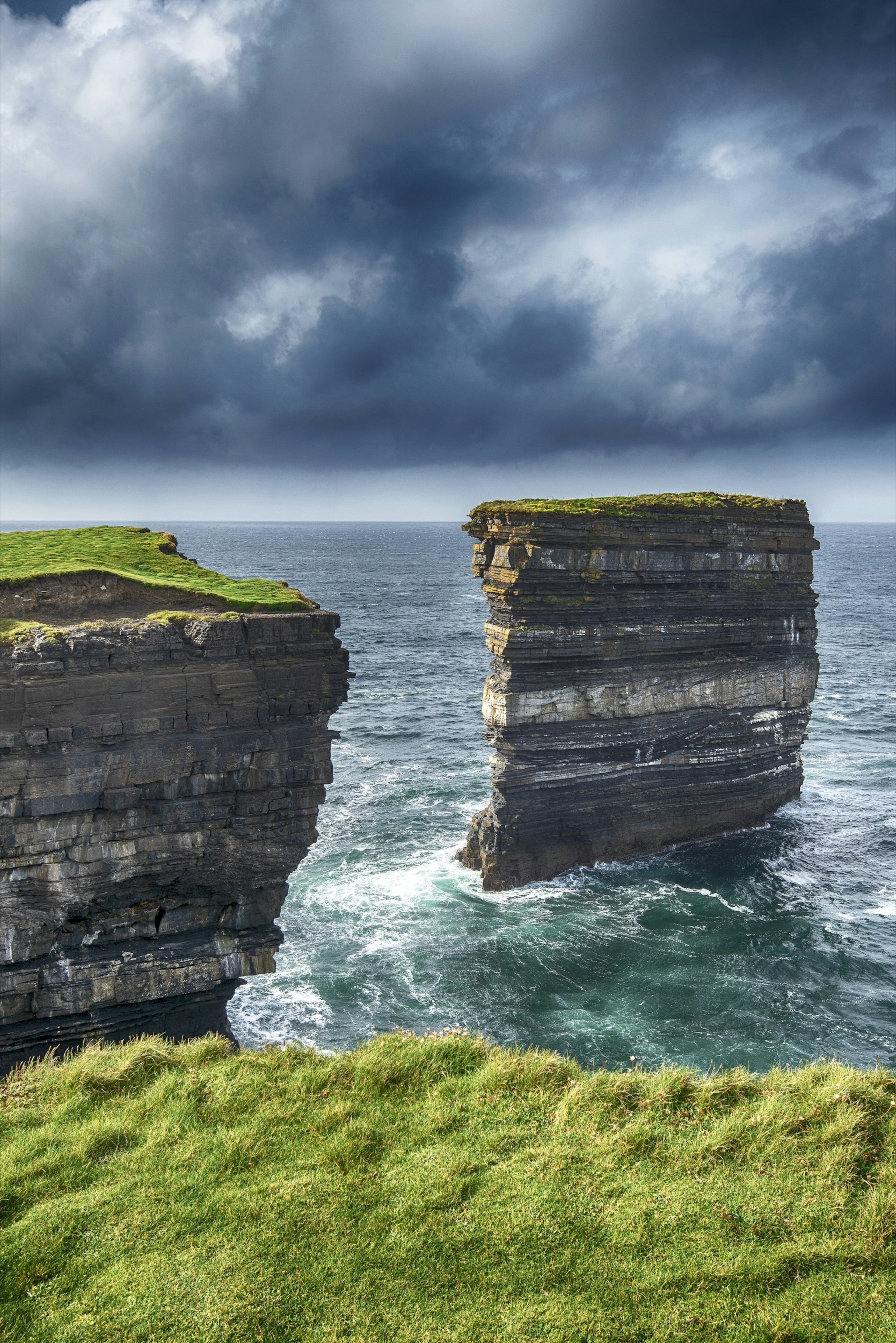 Waves crash at the base of the Dun Briste. There's bright green grass at the top of the natural formation on a dark cloudy day.