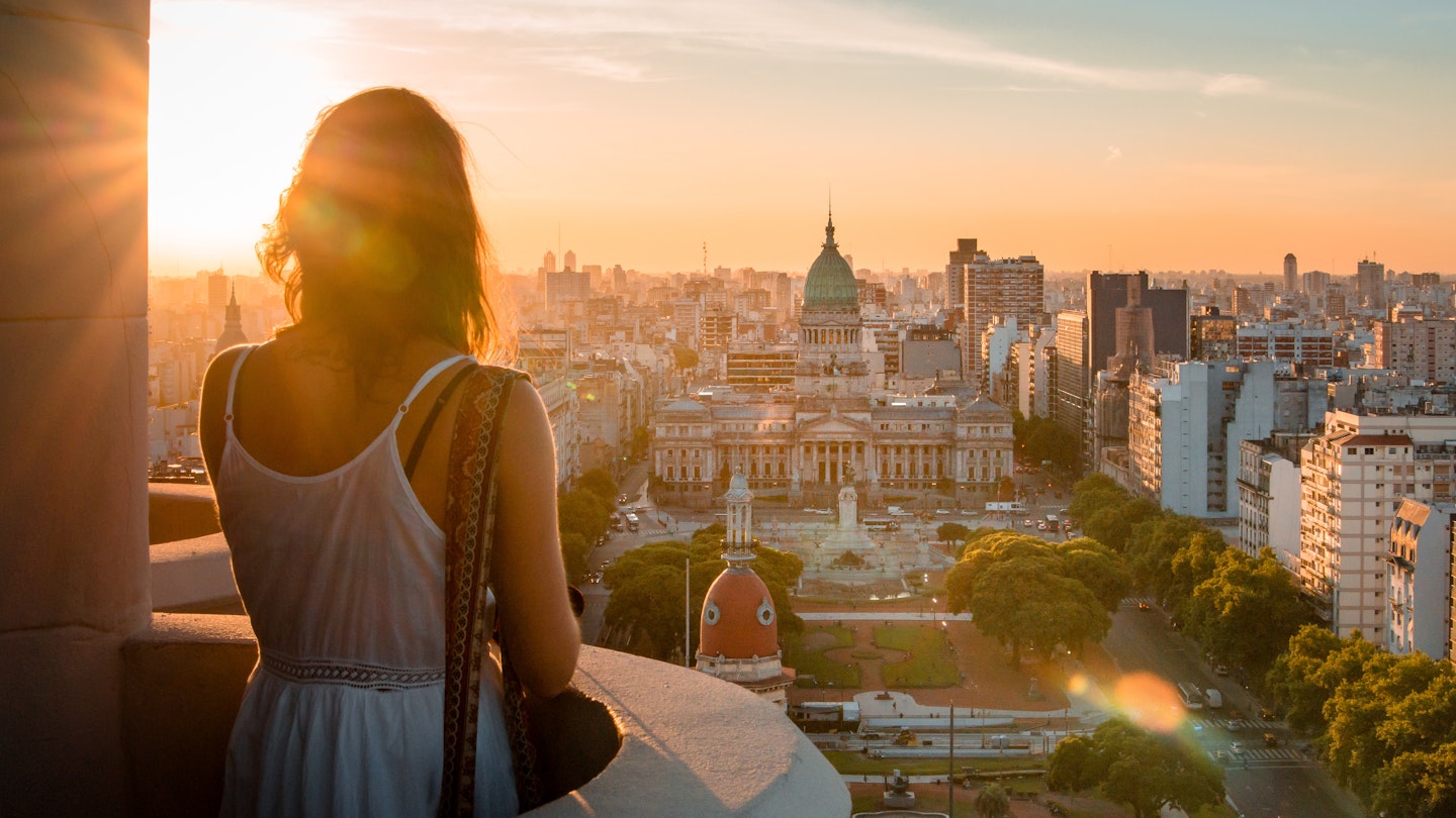 A woman standing on a balcony overlooking the Buenos Aires cityscape during sunset.