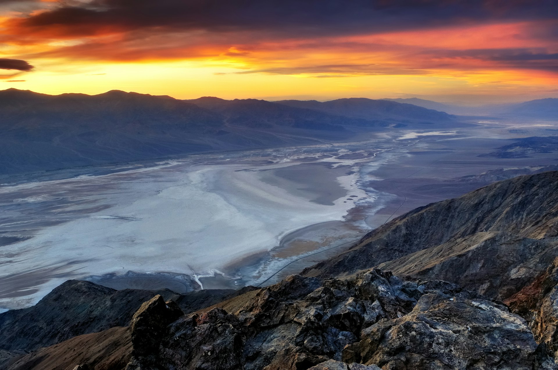 A valley of white salt flats surrounded by gray rocky hills and mountains. The sun is setting in the distance 