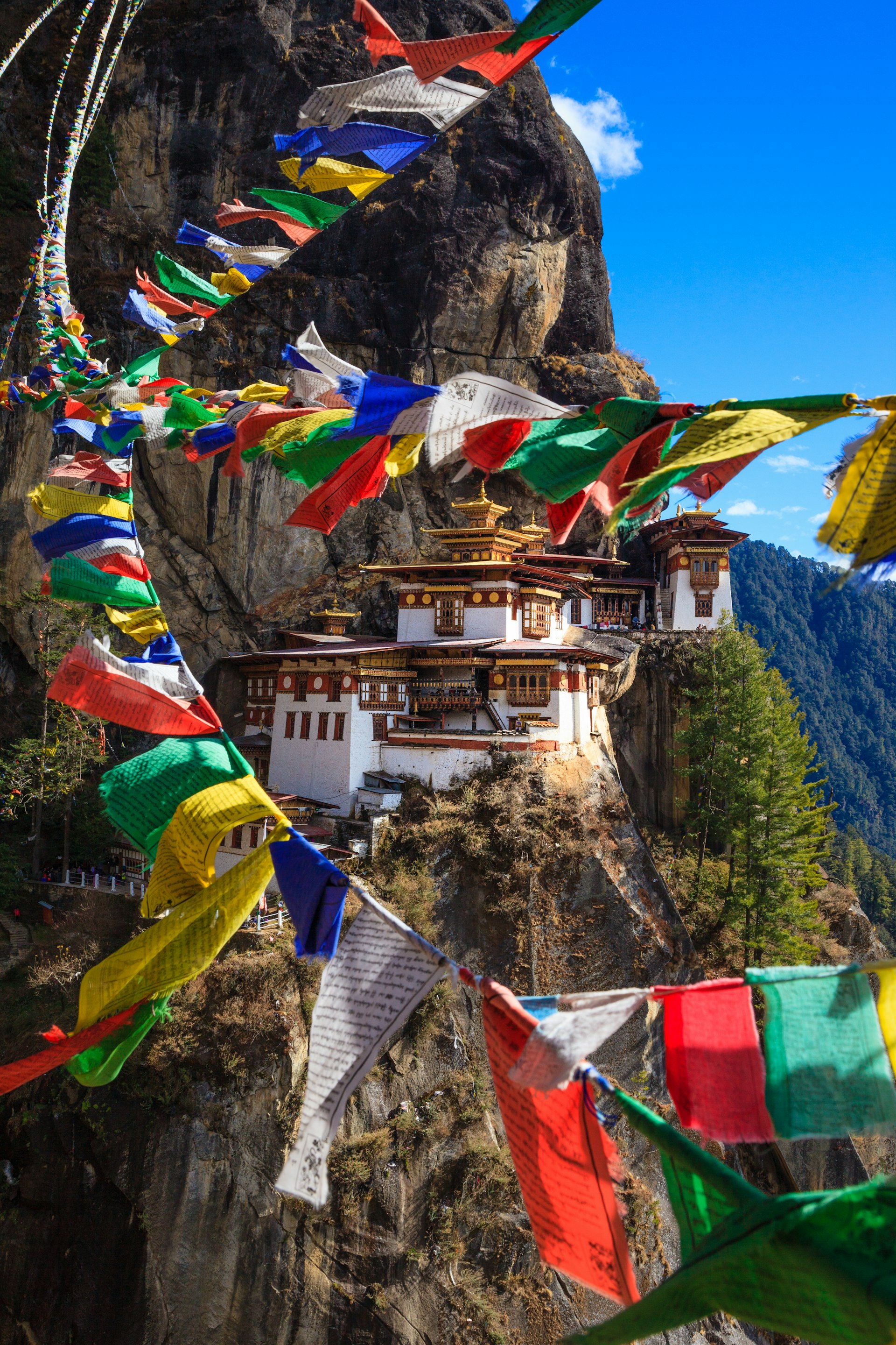 Colorful prayer flags flutter in the breeze. A white monastery structure, built into the side of the mountain, is in the background