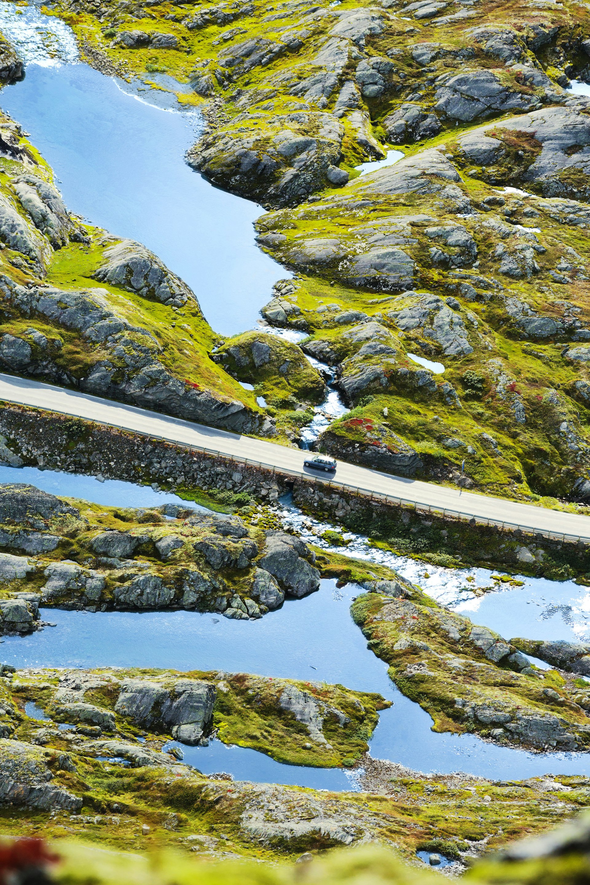 Elevated view of a lone car driving along a road through a fjord with water and rocky outcrops on either side of the raised road