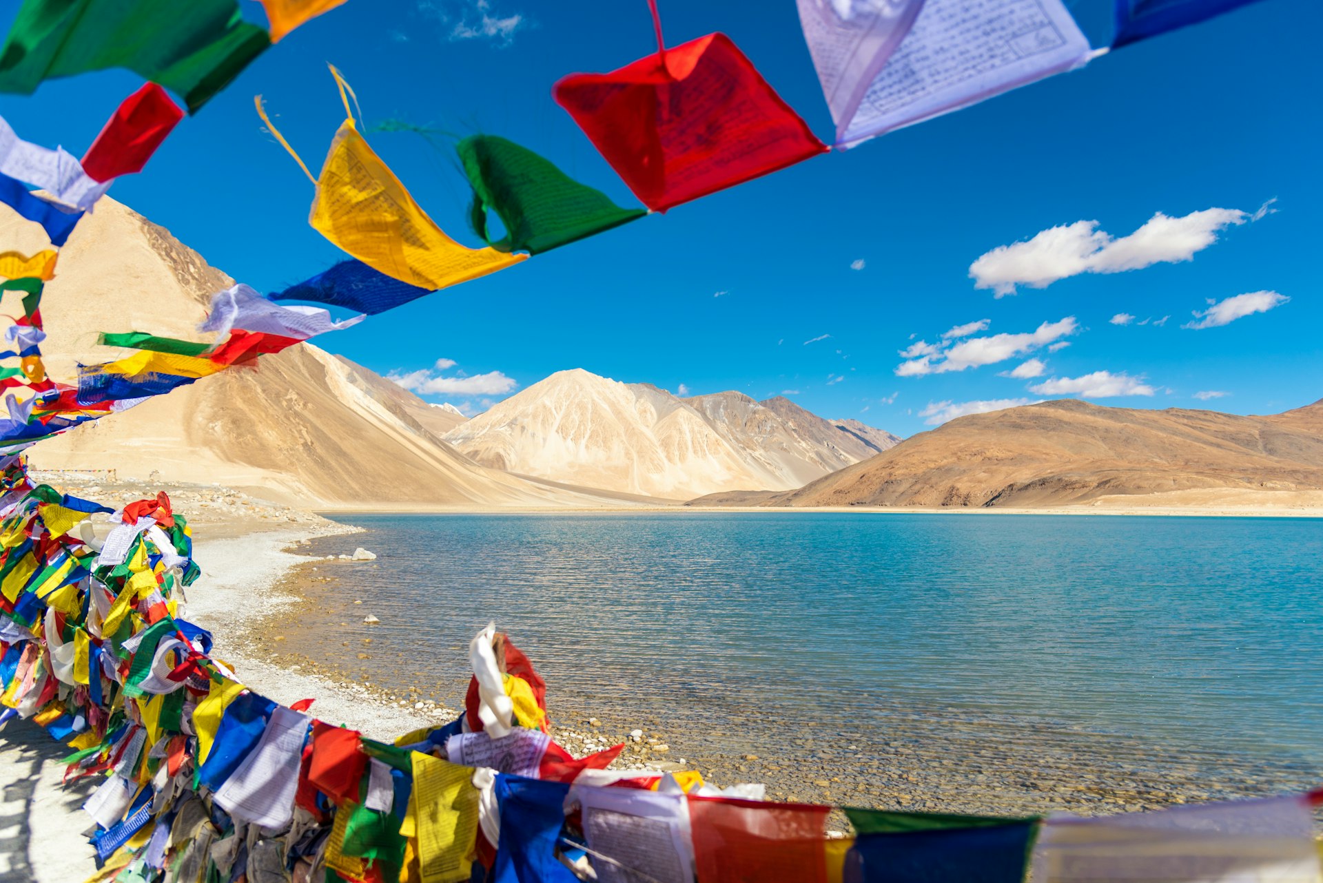 Brightly colored prayer flags flutter in the breeze beside a bright, clear lake surrounded by sandy colored hills