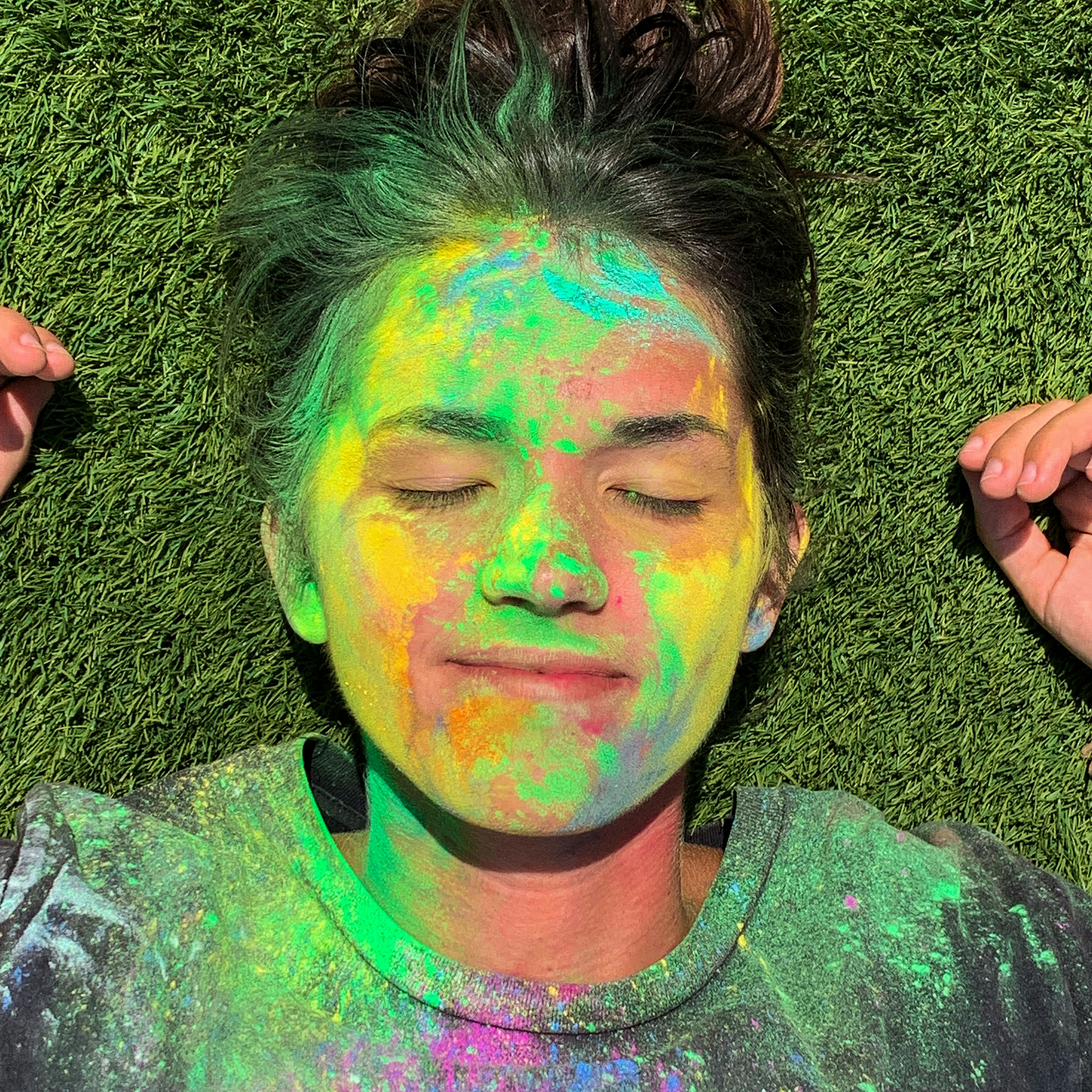 A headshot of a woman lying on grass. Her face is painted in various pastel colors