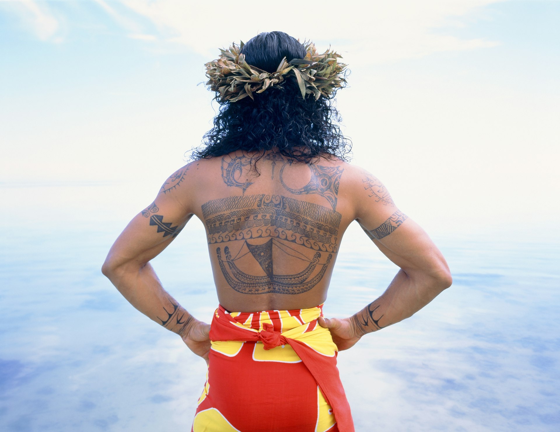 A man with long curly hair wears a floral headdress and a bright yellow and red sarong. His back is covered with traditional Polynesian tattoos.  