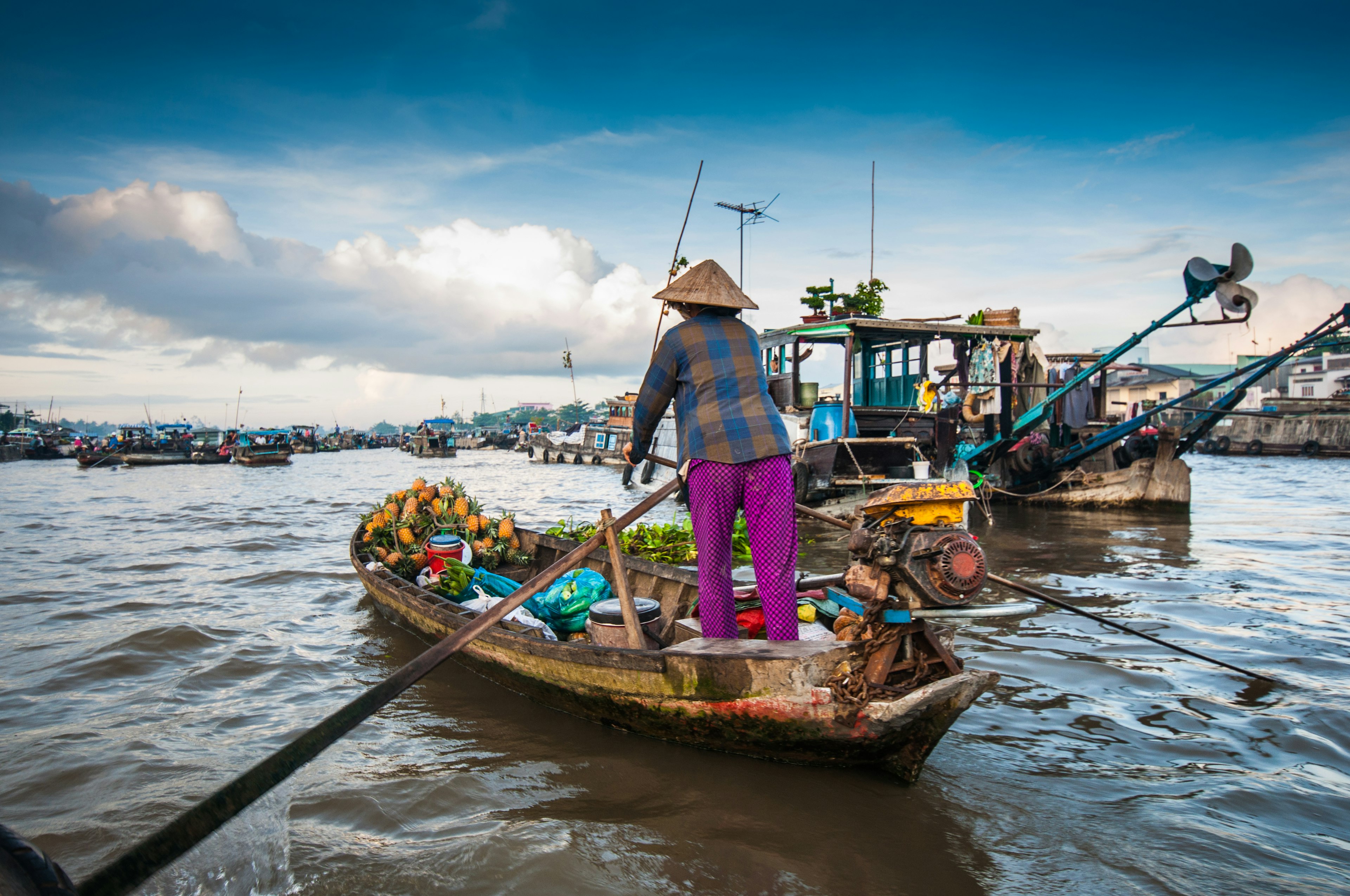 A woman wearing a conical hat pilots a small boat on a wide river