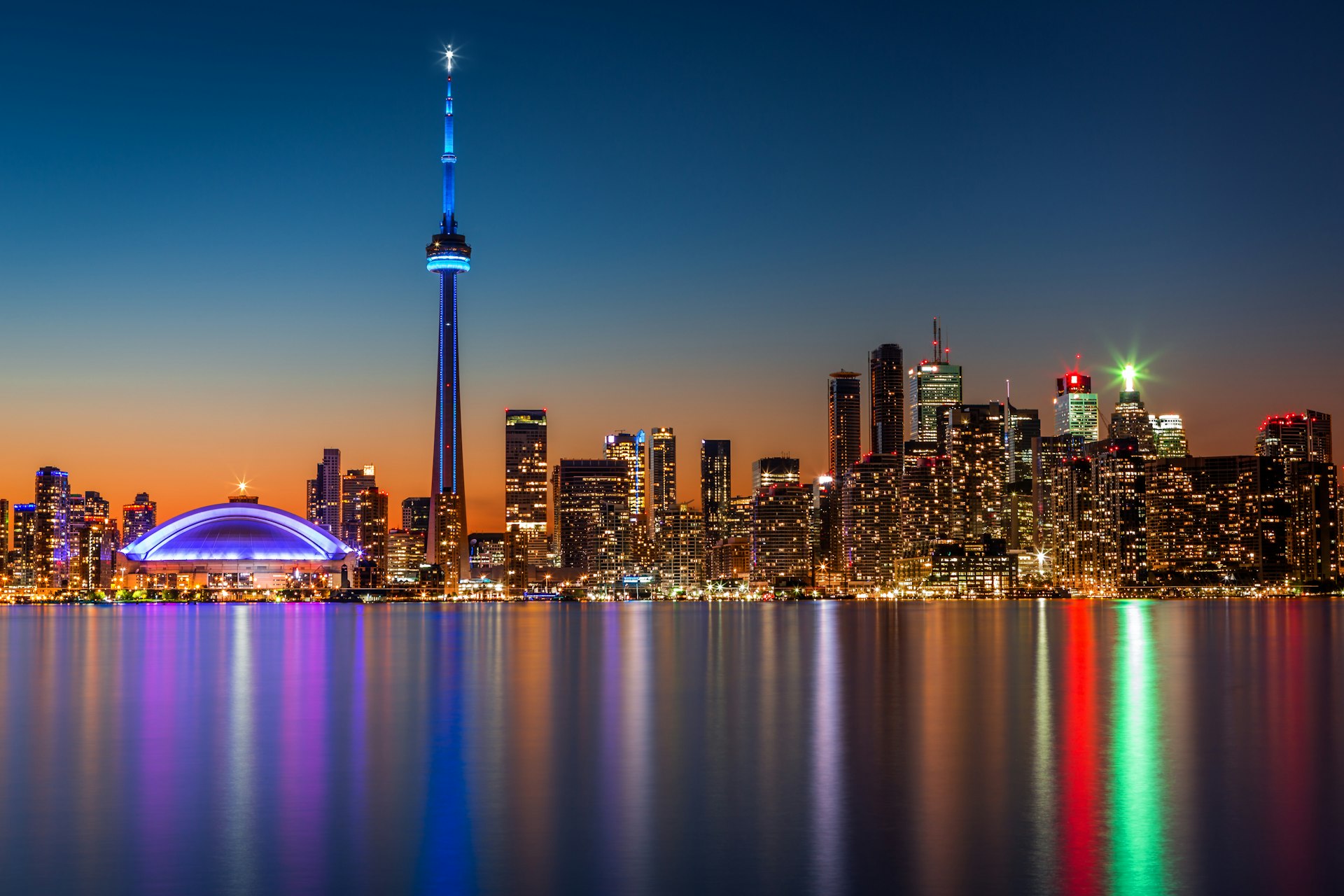 The Toronto skyline at dusk, with a tall needle-like building dominating the shot. Lights are reflected on the water.
