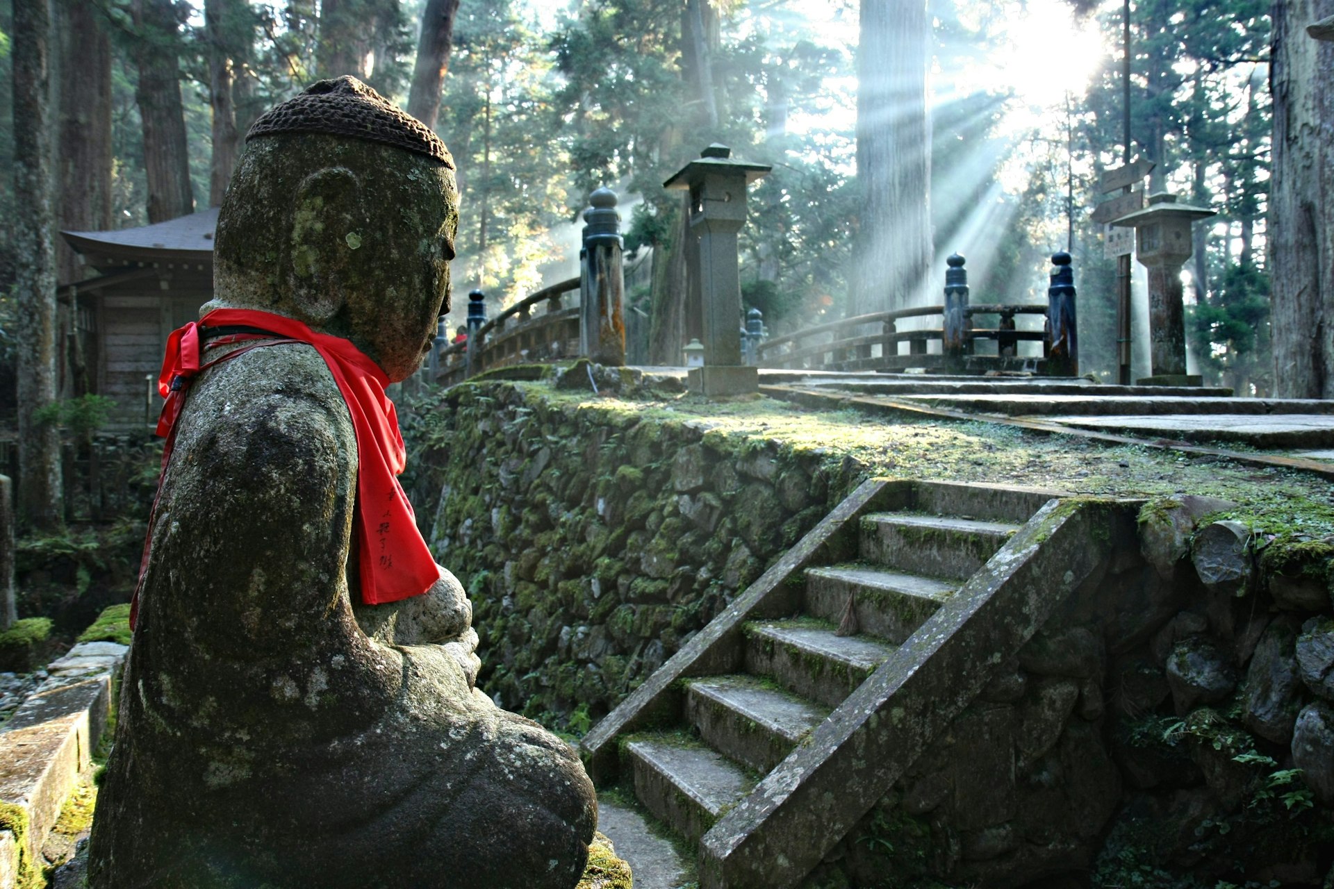 A stone Buddha with a red ribbon around its neck. It is set in forest with beams of sunlight shining between the trees.